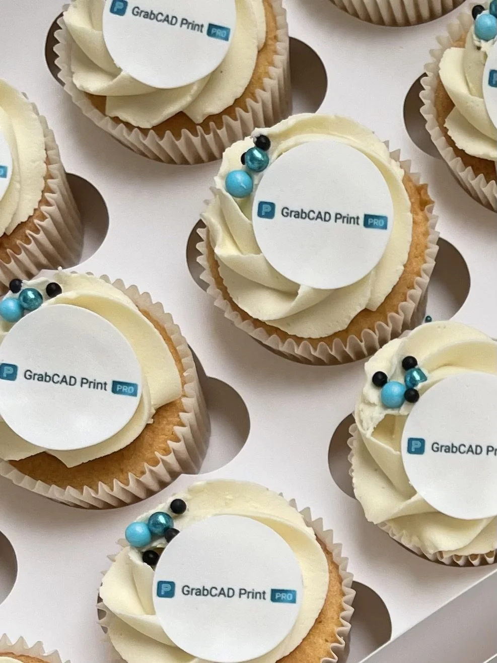Multiple cupcakes decorated with white icing, small decorative blue and black spheres, and edible toppers with the text 'GrabCAD Print PRO' printed on white fondant circles.