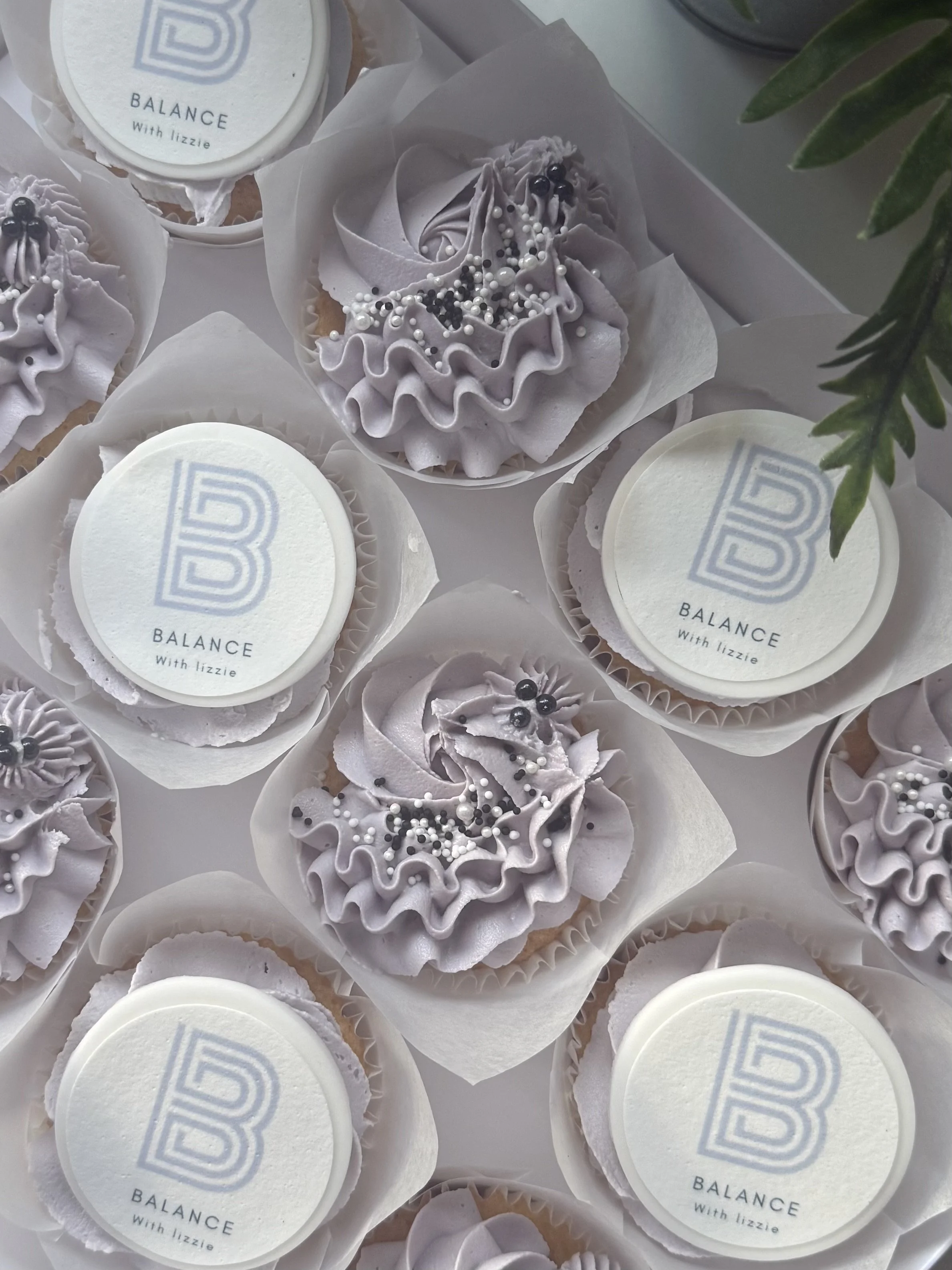 Lavender-colored cupcakes with ruffled frosting and black and white sprinkles, arranged in white cups, accompanied by circular white stickers reading "BALANCE With Lizzie" and a light blue logo.