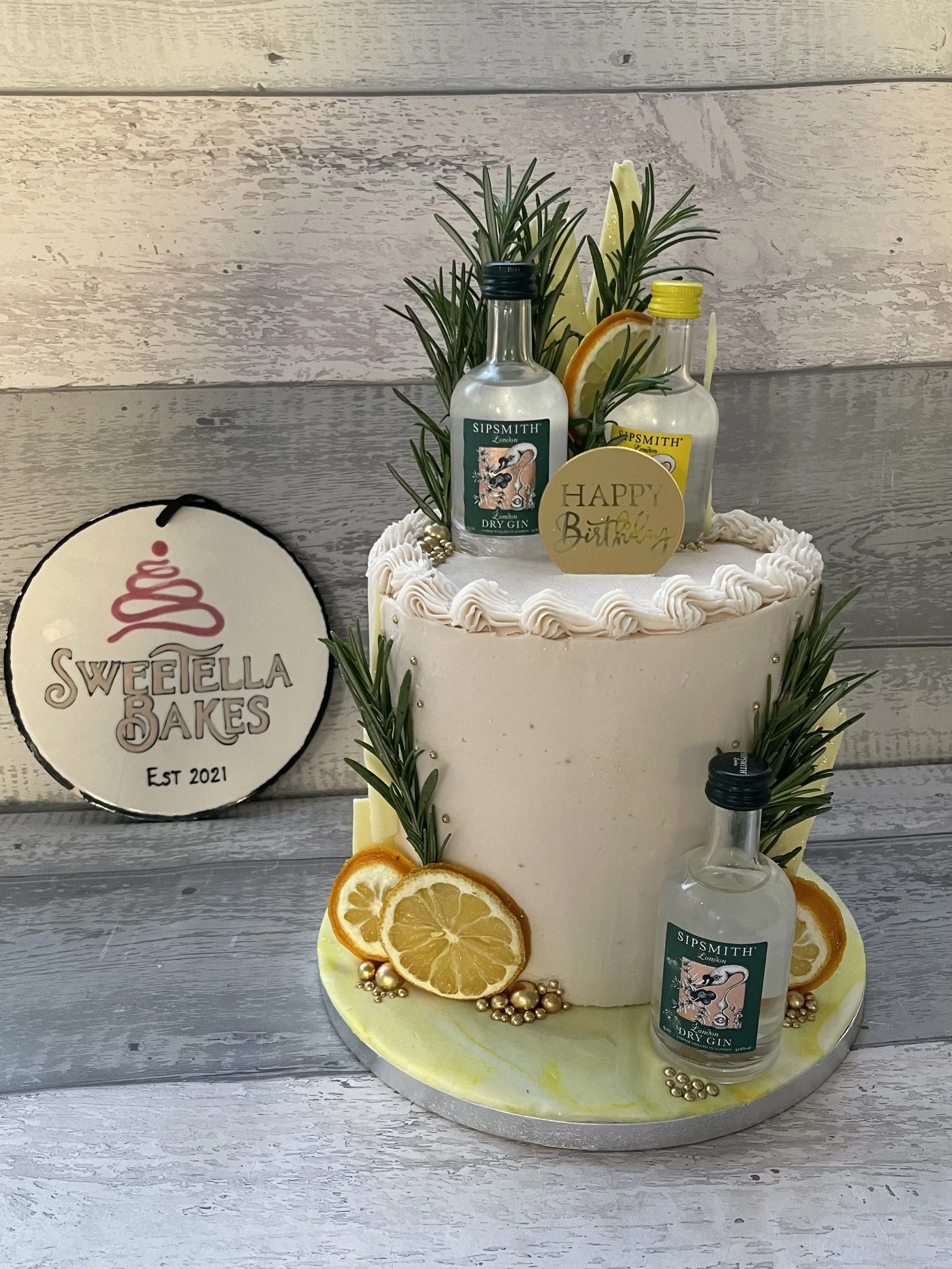 A two-tier white birthday cake decorated with lemon slices, sprigs of greenery, and small gold beads, topped with miniature gin bottles, lemon and orange slices, and a gold 'Happy Birthday' sign.