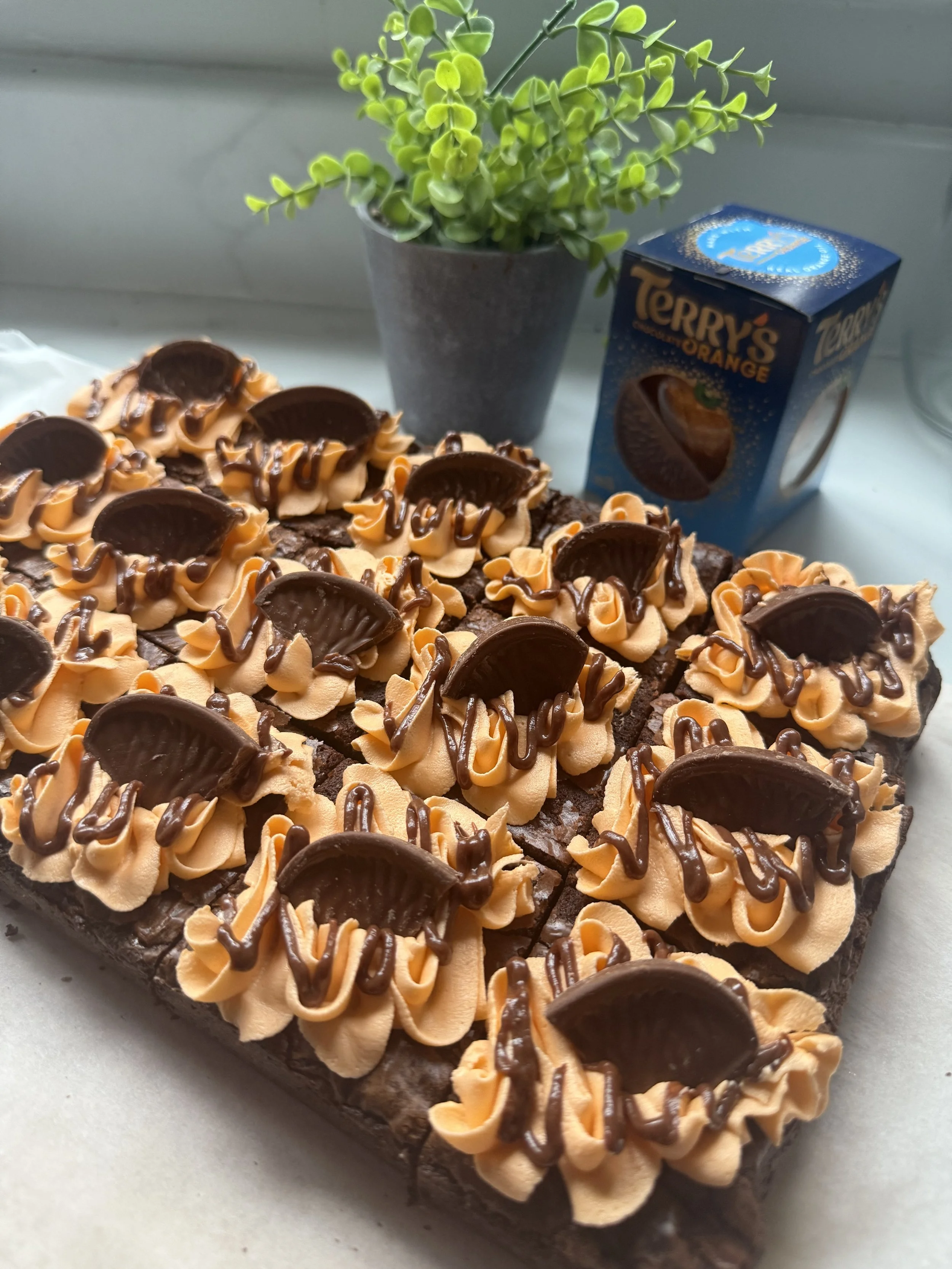 Chocolate brownie decorated with swirls of orange frosting and topped with Halves of Reese's Peanut Butter Cups, with a potted green plant and a box of Terry's Orange chocolate in the background.