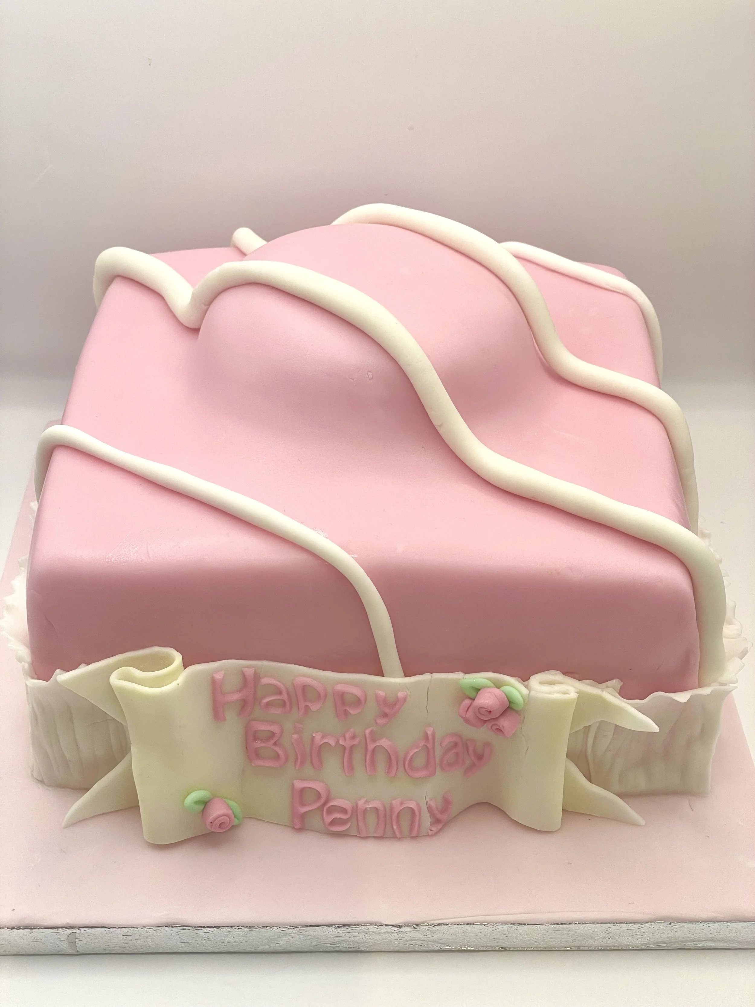 Pink birthday cake decorated with white icing squiggles and a white banner with pink letters that reads 'Happy Birthday Penny' with small pink roses and green leaves.