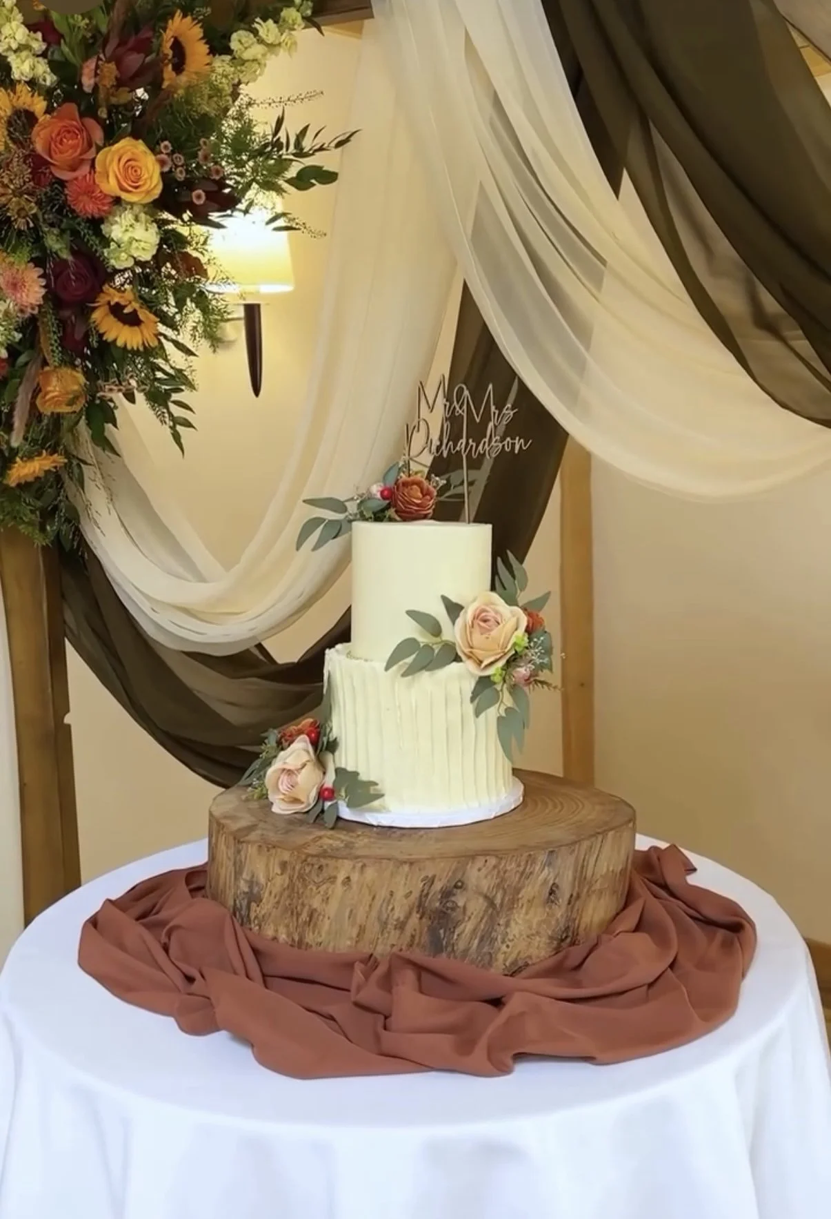 A wedding cake with two tiers, decorated with pink and peach flowers and green leaves, sitting on a wooden base with a brown cloth draped underneath, against a backdrop of white and dark brown drapes and a floral arrangement.