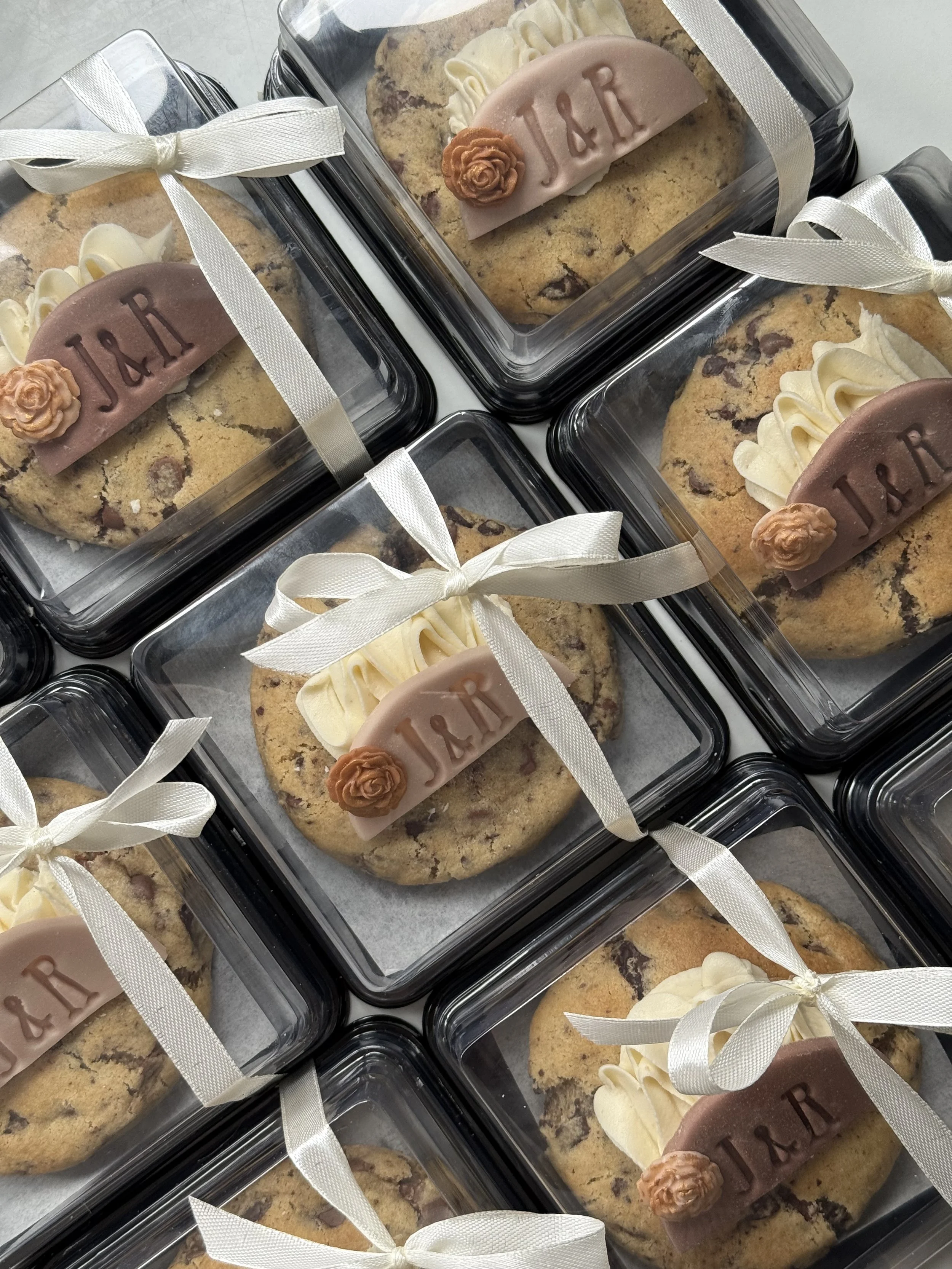Several cookies decorated with white icing, cream, and a pink fondant label with the initials 'J&R' and a small fondant flower, all packaged in clear plastic boxes and tied with white ribbons.