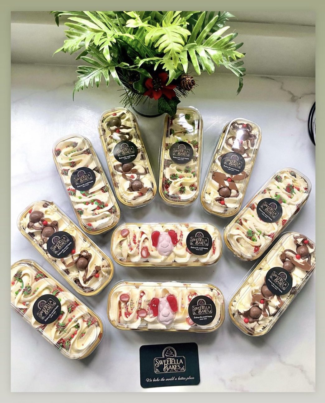 Various decorated cakes from Sweella Bakes arranged in a circle with a poinsettia and green leaves centerpiece, on a white surface.