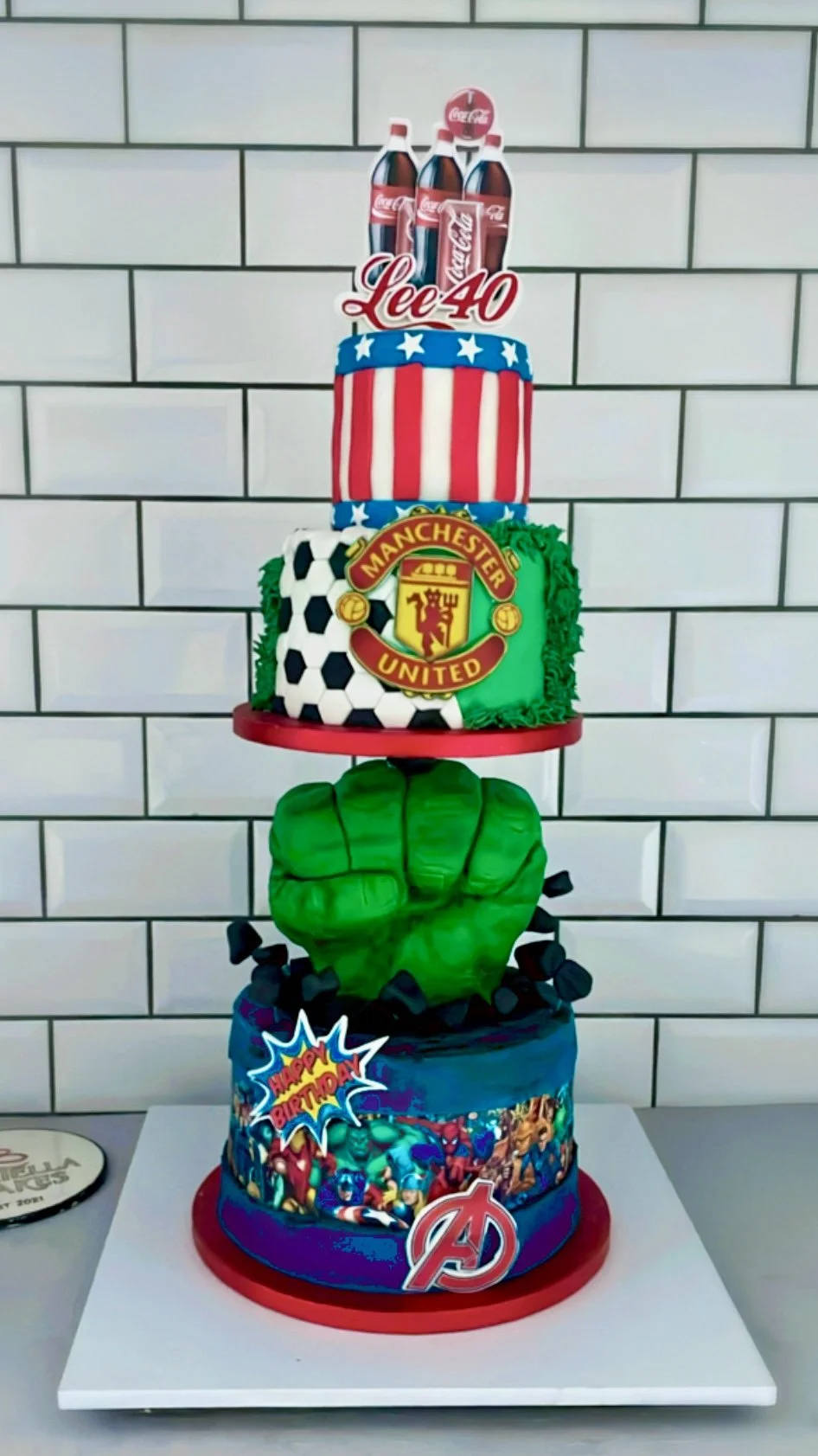 Multi-tiered birthday cake featuring Coca-Cola, Manchester United, Marvel superheroes, and Avengers themes, topped with a Coca-Cola logo, Manchester United crest, and Marvel characters.