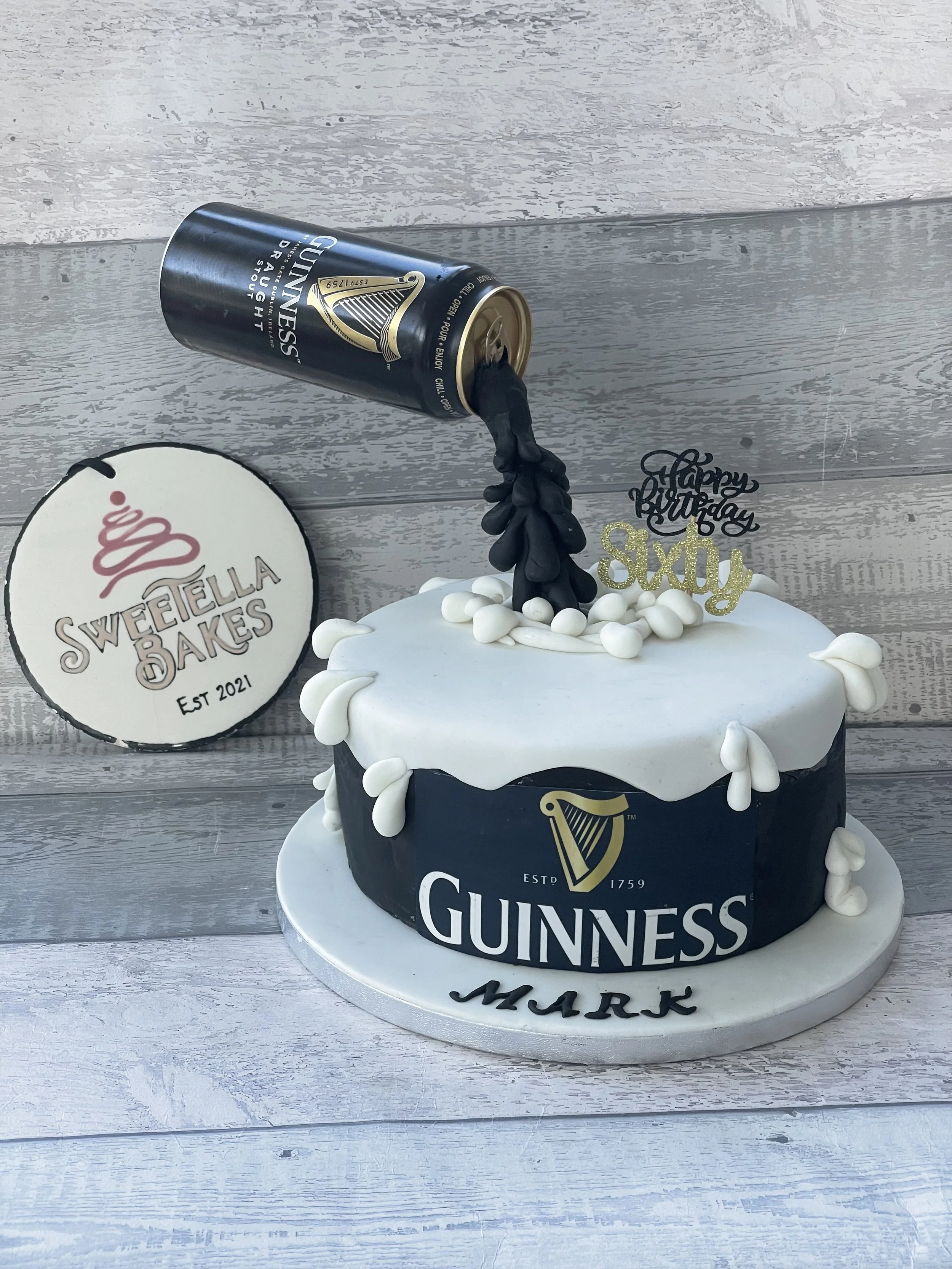 A Guinness-themed birthday cake with a black and white color scheme. It has a gold Guinness logo, white icing decorations, and a black candle shaped like a Guinness tap pouring into the cake. There are also cake toppers reading "Happy Birthday" and "