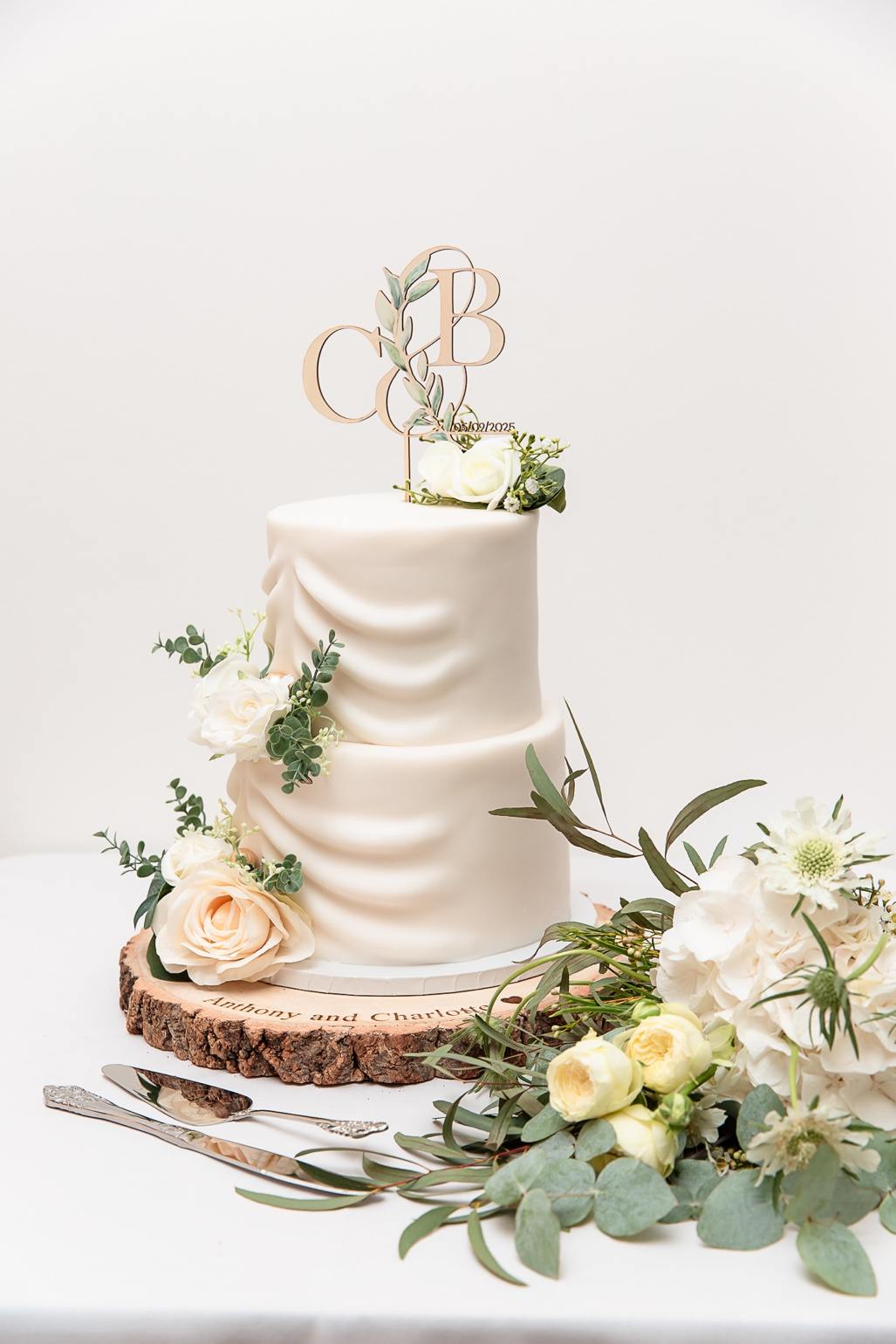 Two-tier white wedding cake decorated with white roses, green leaves, and a custom cake topper with initials 'C' and 'B'. The cake sits on a wooden slab with engraved text, surrounded by additional flowers and greenery, and a cake knife and server in