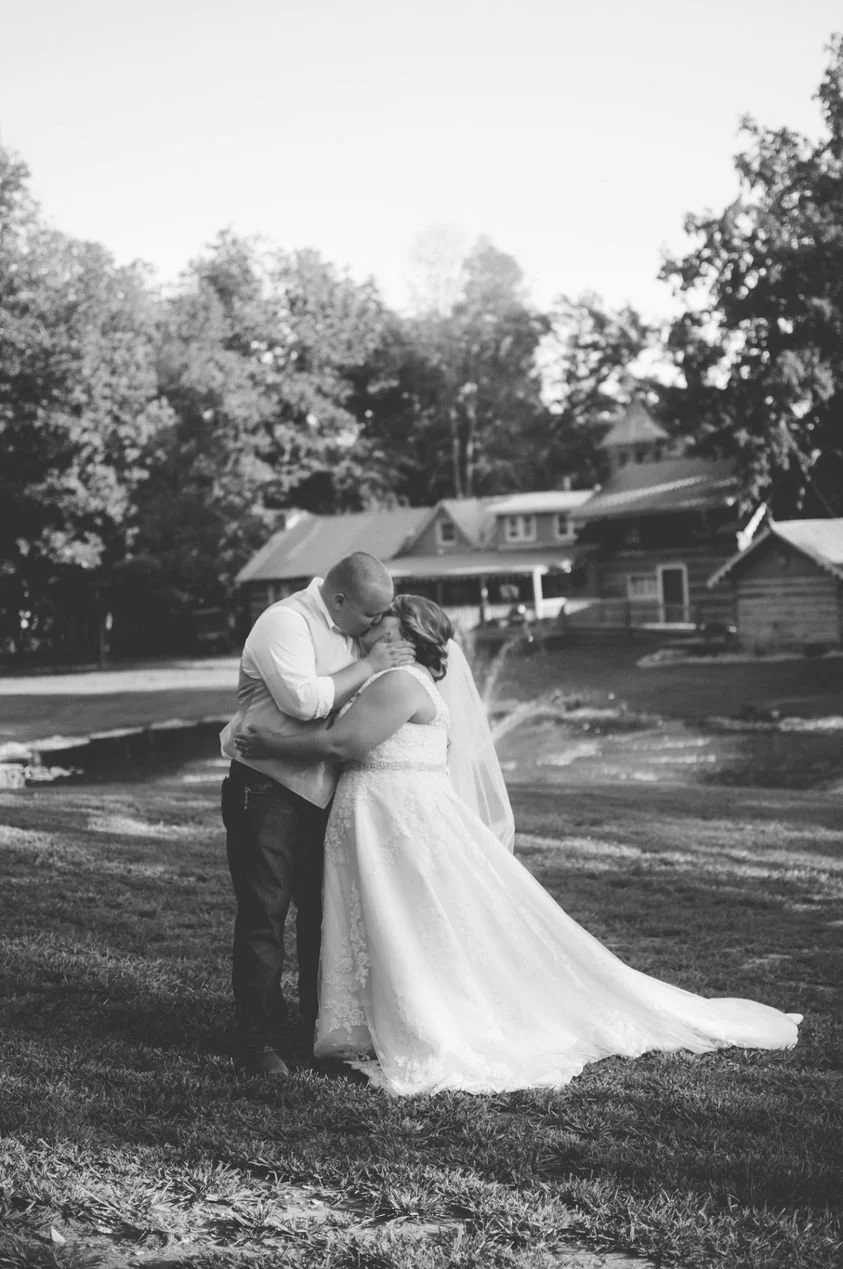 A couple in wedding attire sharing a kiss outdoors on a lawn with trees and house in the background, black and white photo.