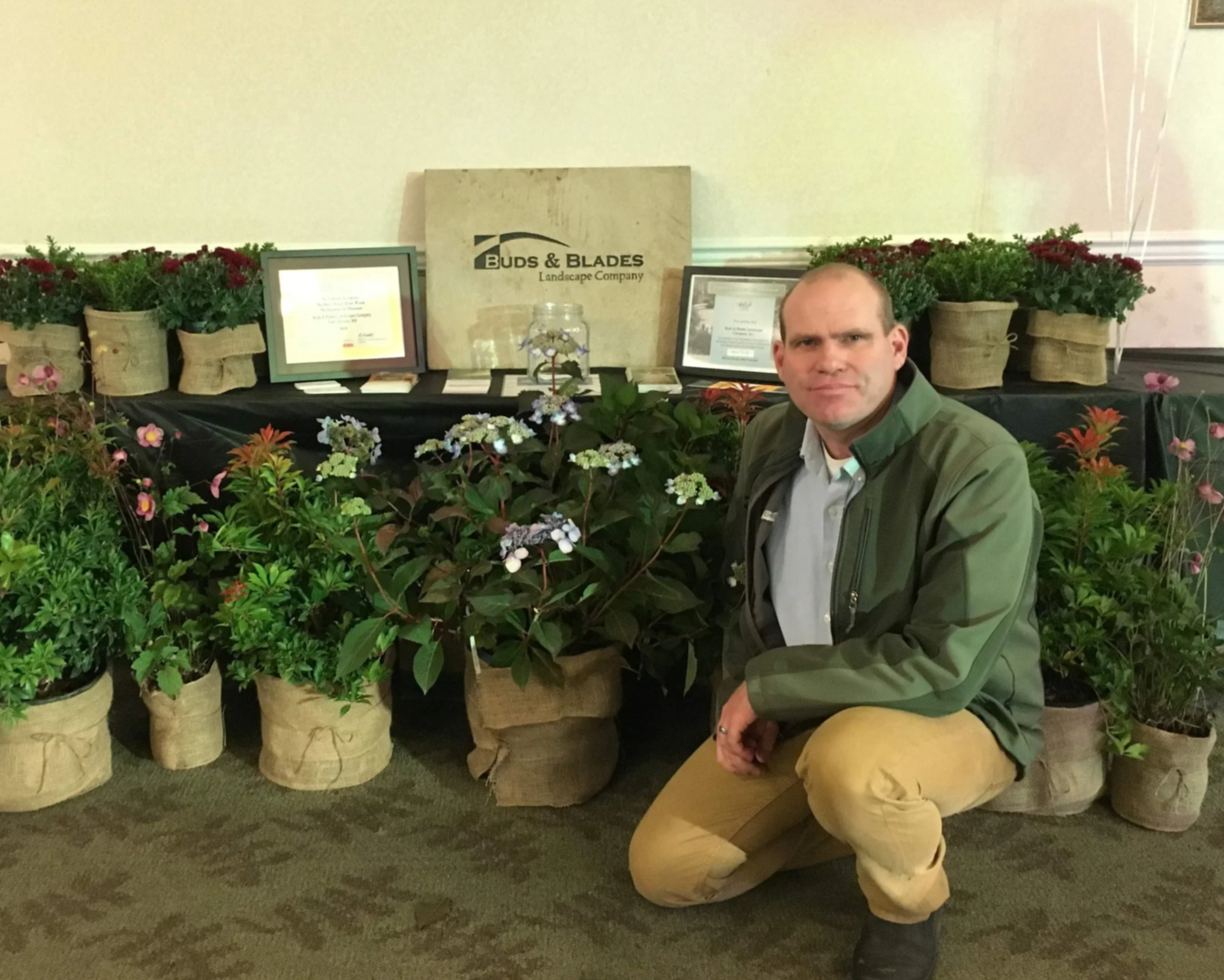 Rick Longnecker crouching next to potted plants at a landscaping display, with certificates and a sign reading 'Buds & Blades' in the background.
