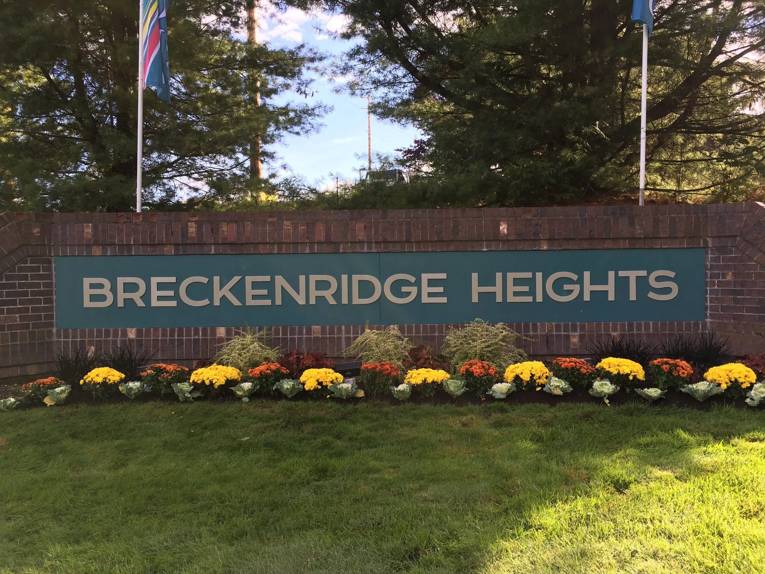 Sign for Breckenridge Heights with flower bed in front and flagpoles with flags behind, surrounded by trees.