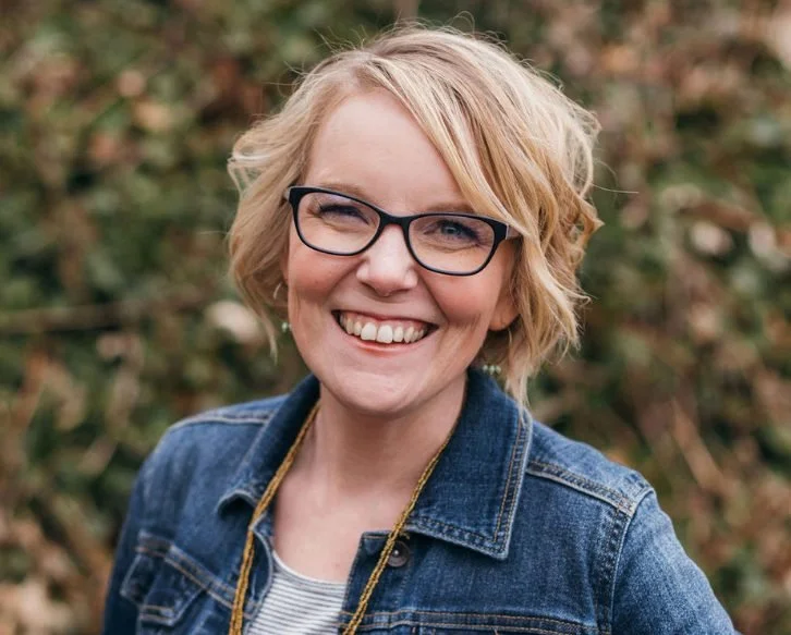 A smiling woman with short blonde hair, glasses, wearing a denim jacket outdoors.
