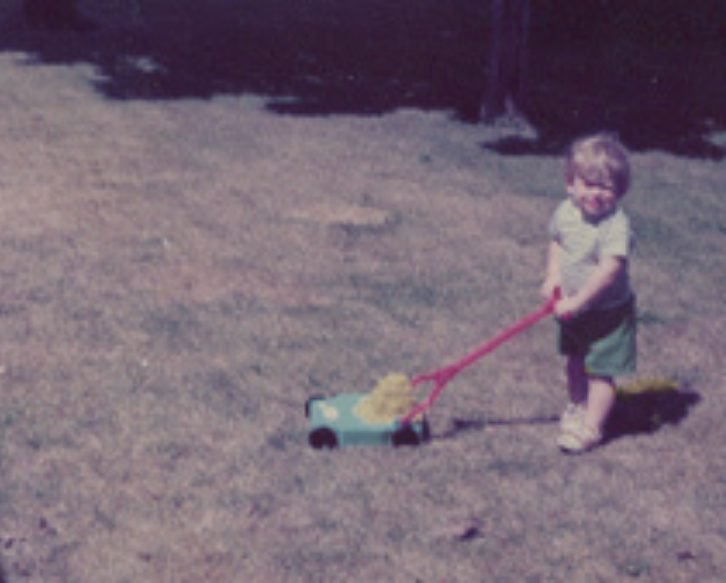 Rick Longnecker as a young child pulling a toy lawnmower with a yellow object inside on a grassy yard.