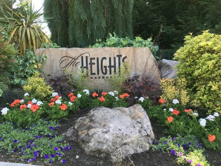 A landscaped garden with colorful flowers, green shrubs, and a large rock in front of a stone sign that reads 'The EIGHT.'