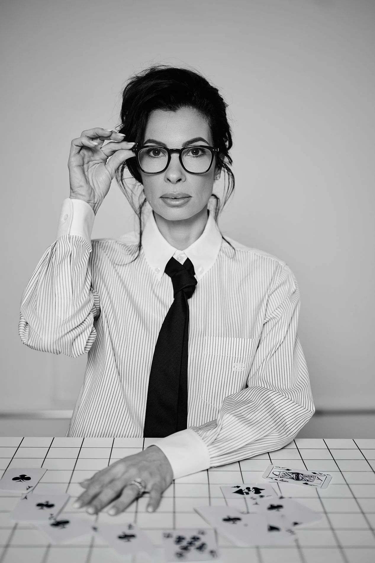 Black and white photo of a woman with dark hair and glasses, wearing a striped shirt and a tie, sitting at a table with playing cards, with her hand reaching out and the other adjusting her glasses.