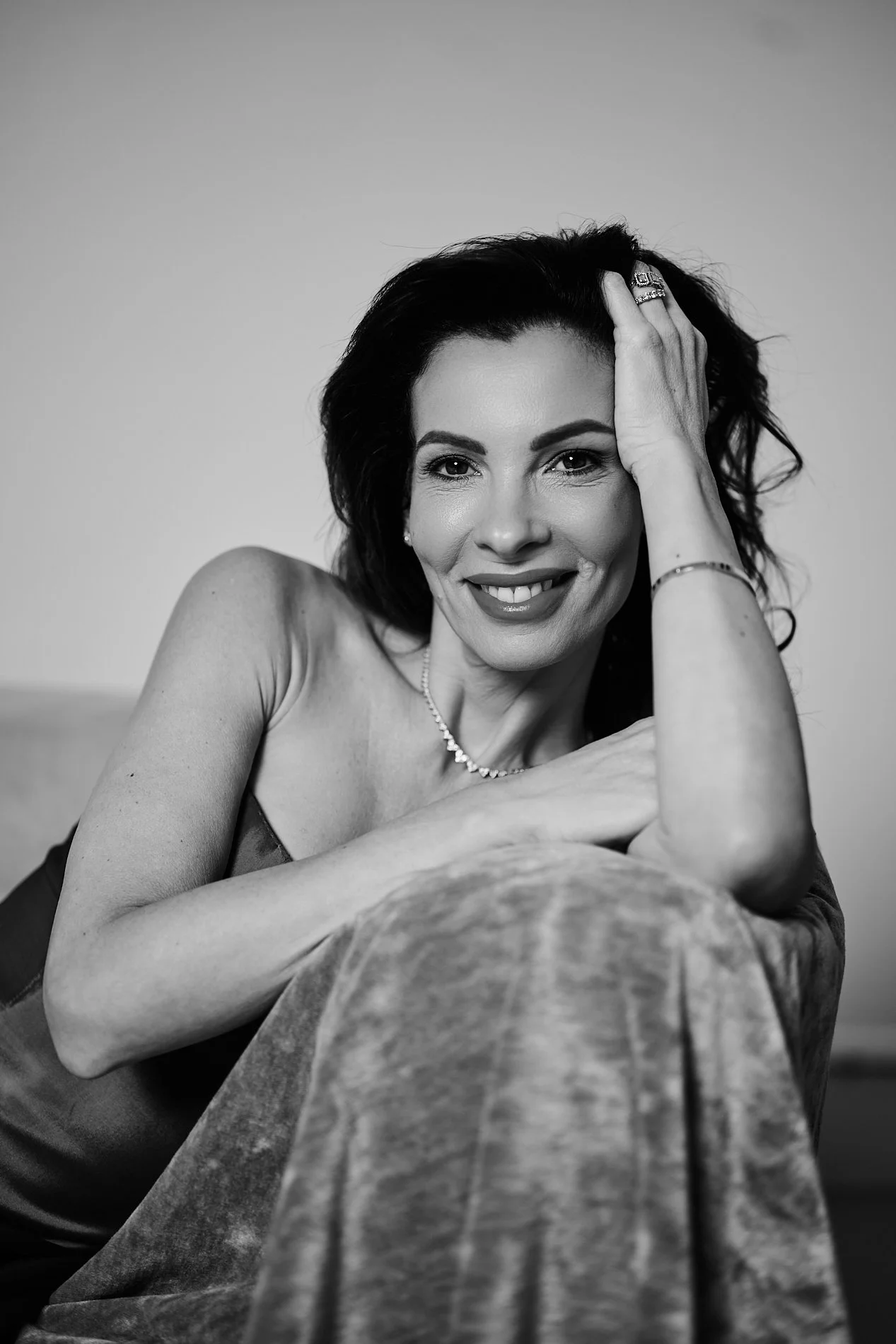 Black and white portrait of a woman with dark hair, smiling, with her head resting on her hand, wearing jewelry and a sleeveless top.