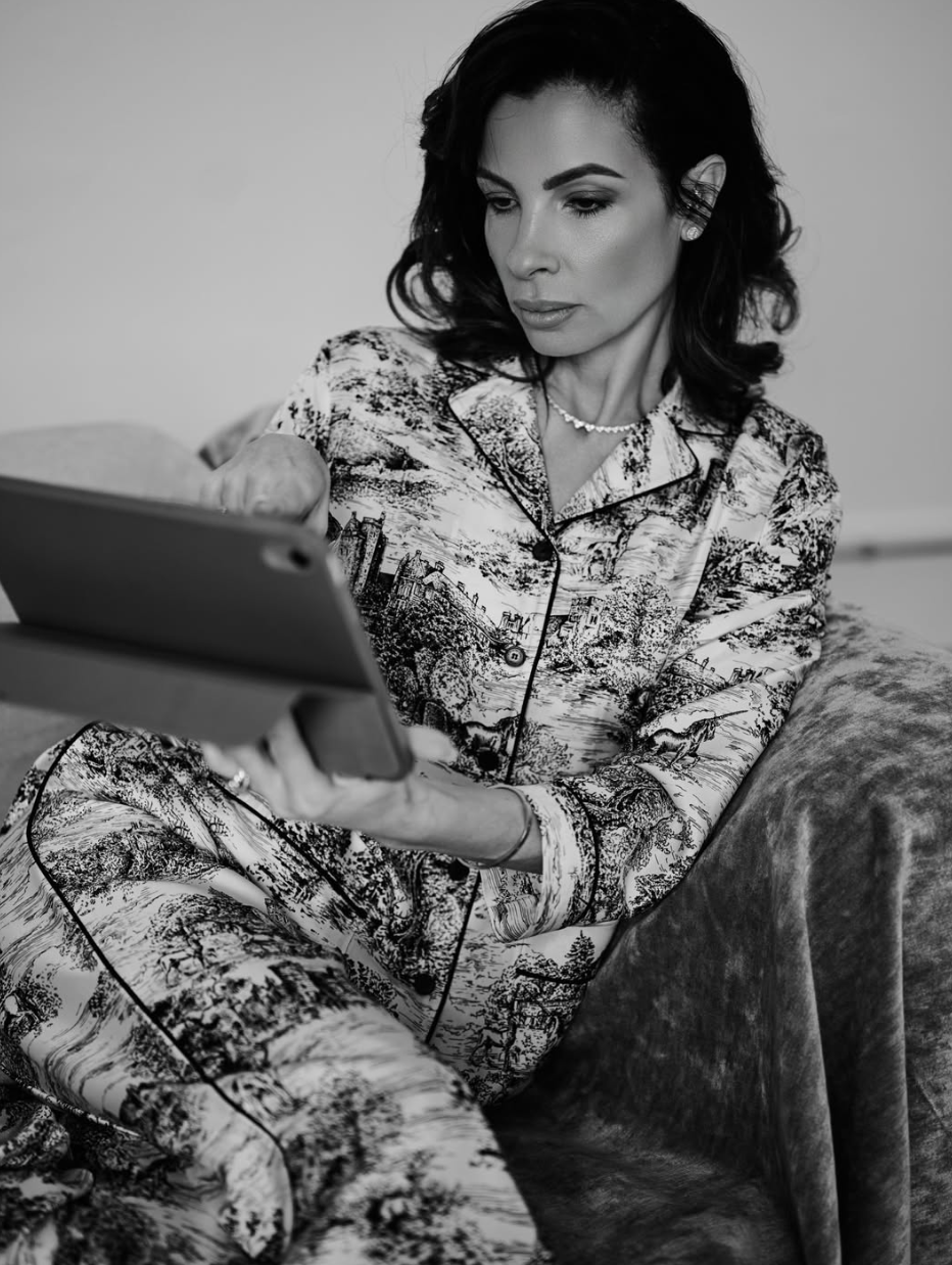 A woman with dark wavy hair checking her phone while sitting on a bed. She is wearing patterned pajamas with a scenic landscape design, a pearl necklace, and earrings.