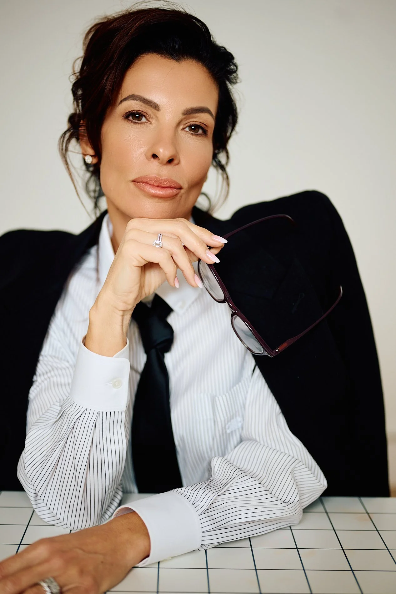A woman with dark hair styled in loose waves, wearing a black blazer over a white striped shirt with a black tie, sitting at a tiled table, holding sunglasses, and looking confidently at the camera.