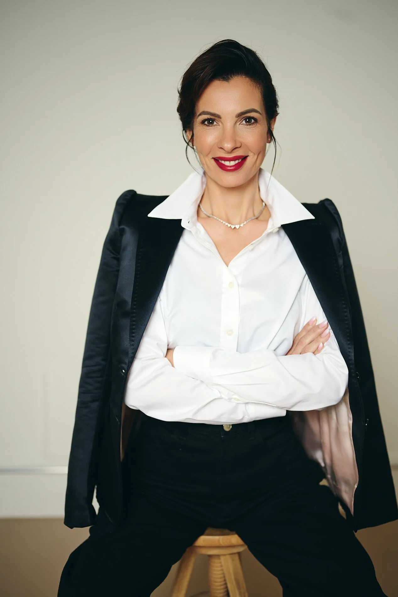 A woman with dark hair styled back, wearing a white button-up shirt, black blazer draped over her shoulders, and black pants, sitting on a stool against a neutral background, smiling with arms crossed.