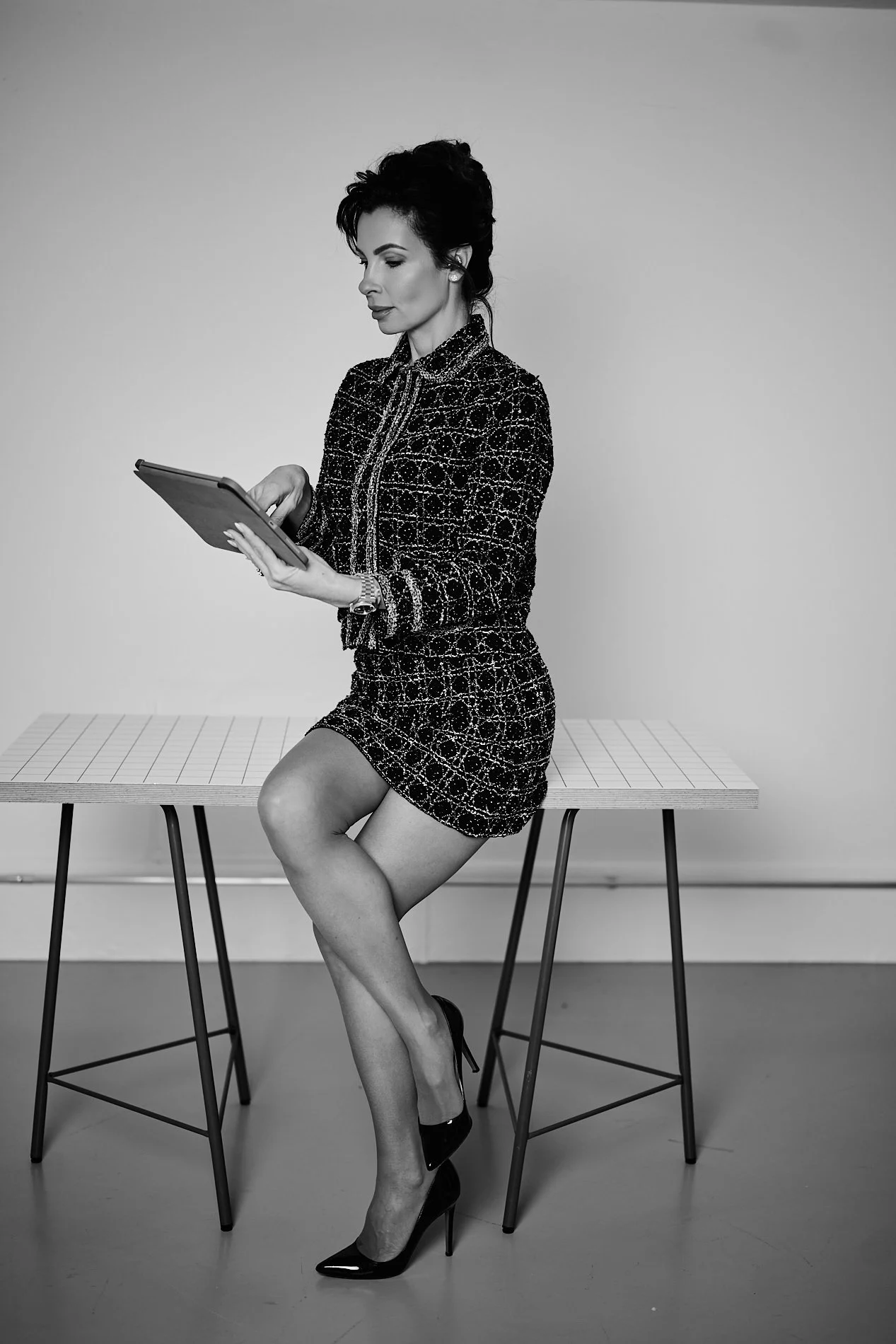 Black and white photo of a woman sitting on a table, holding a tablet, dressed in a patterned blazer and skirt, wearing high heels.
