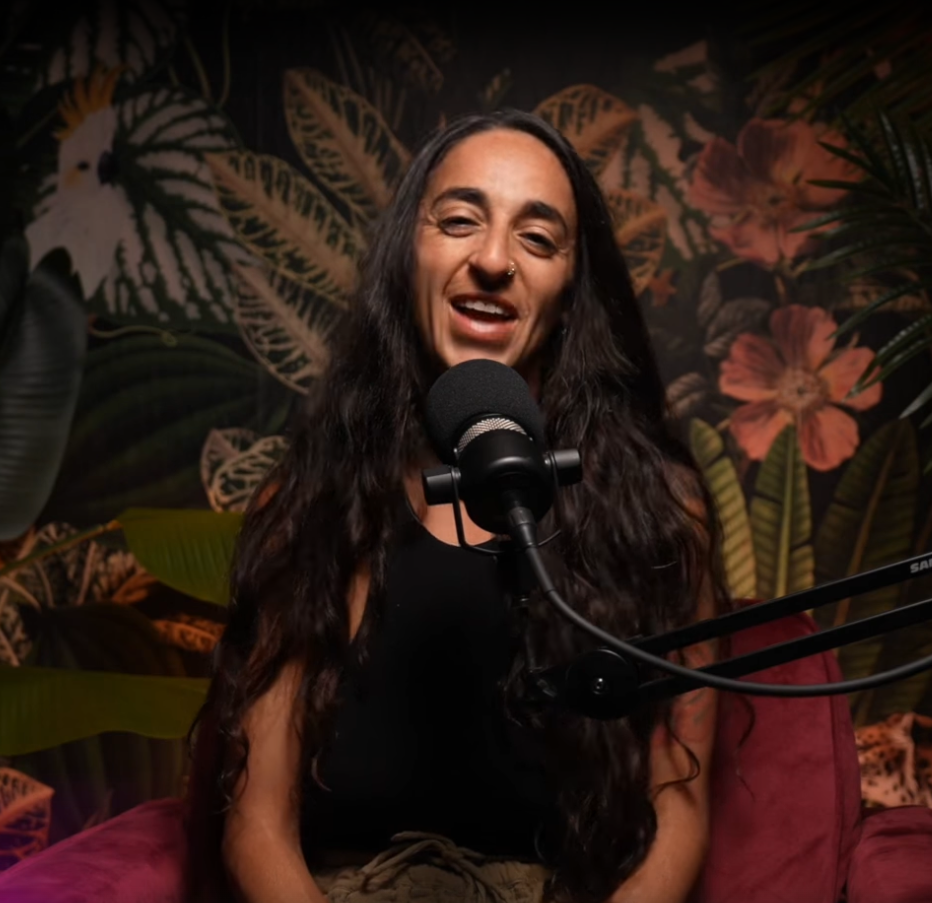 A woman with long dark hair, a nose piercing, and wearing a black top, is speaking into a microphone in front of a tropical plant-themed backdrop.