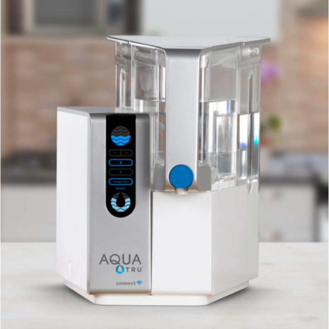 A white countertop water filtration machine with digital controls and a transparent water chamber, labeled "AQUA TRU."