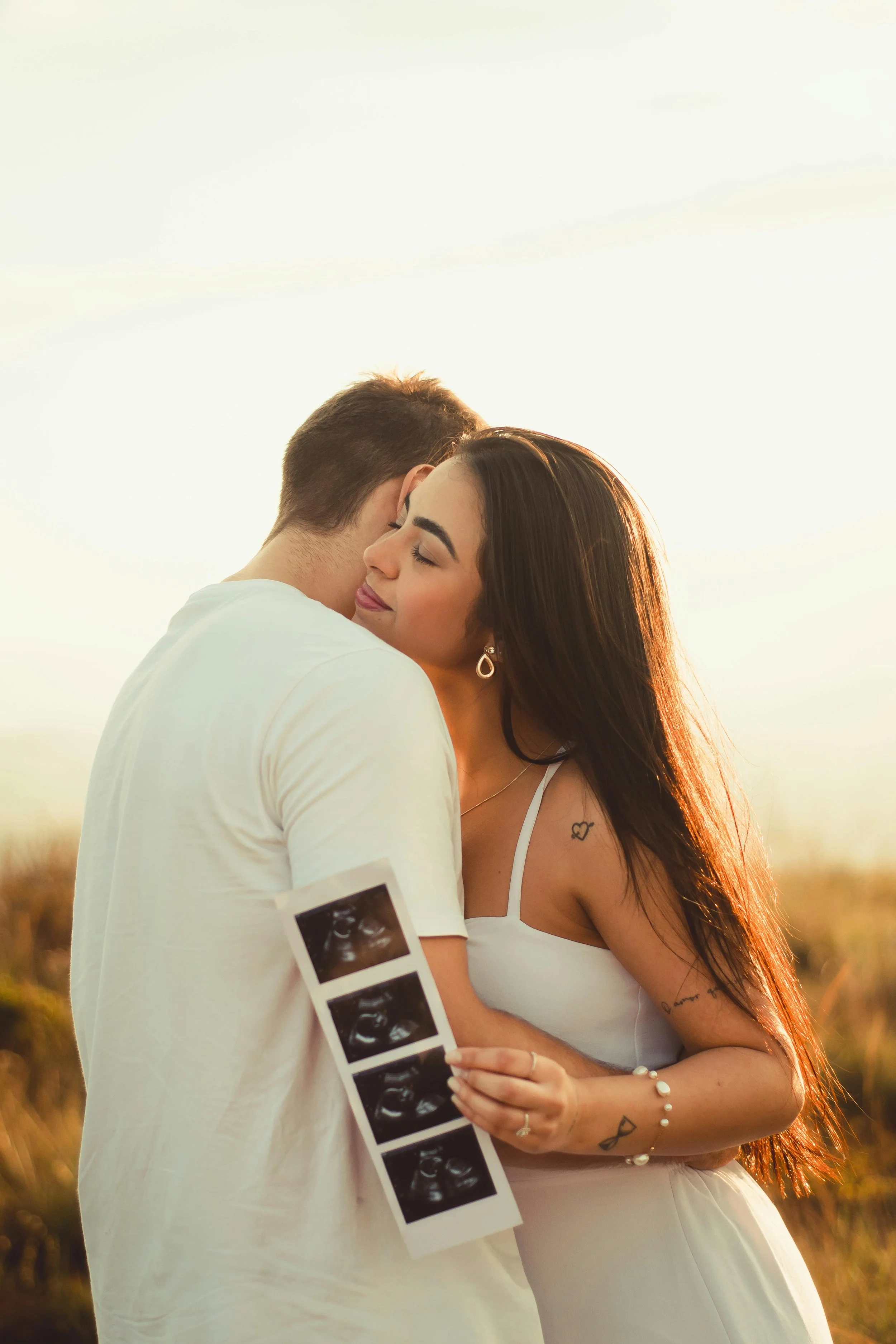 A couple embracing outdoors during sunset, with the woman holding ultrasound images, suggesting a pregnancy announcement.
