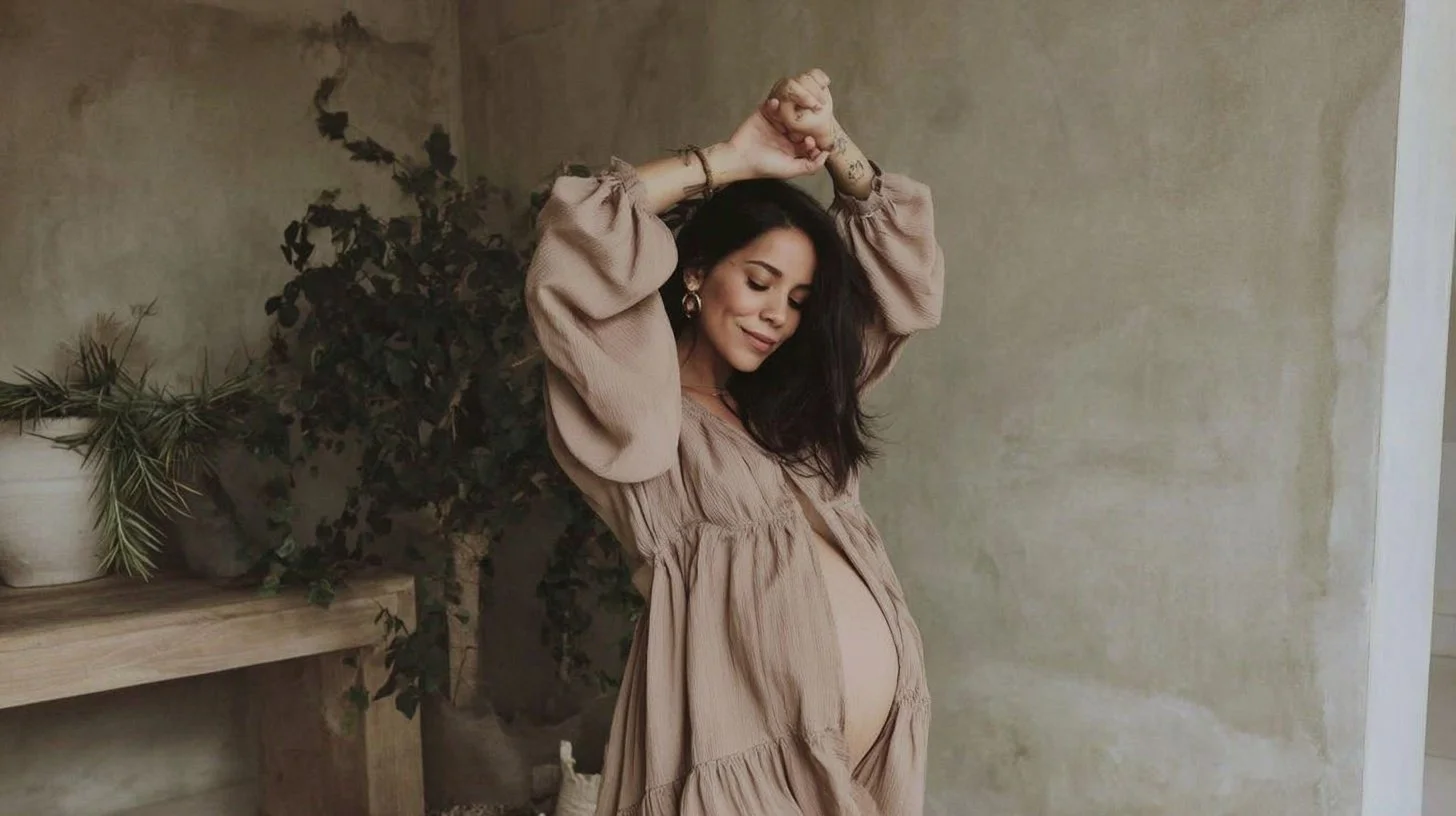 Pregnant woman with long dark hair wearing a beige dress, standing against a textured, neutral-colored wall with greenery on a wooden table, smiling with eyes closed and hands raised above her head.