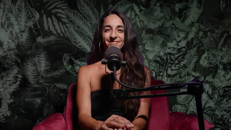 Woman with long dark hair sitting in front of a microphone, smiling, with leafy wallpaper background.