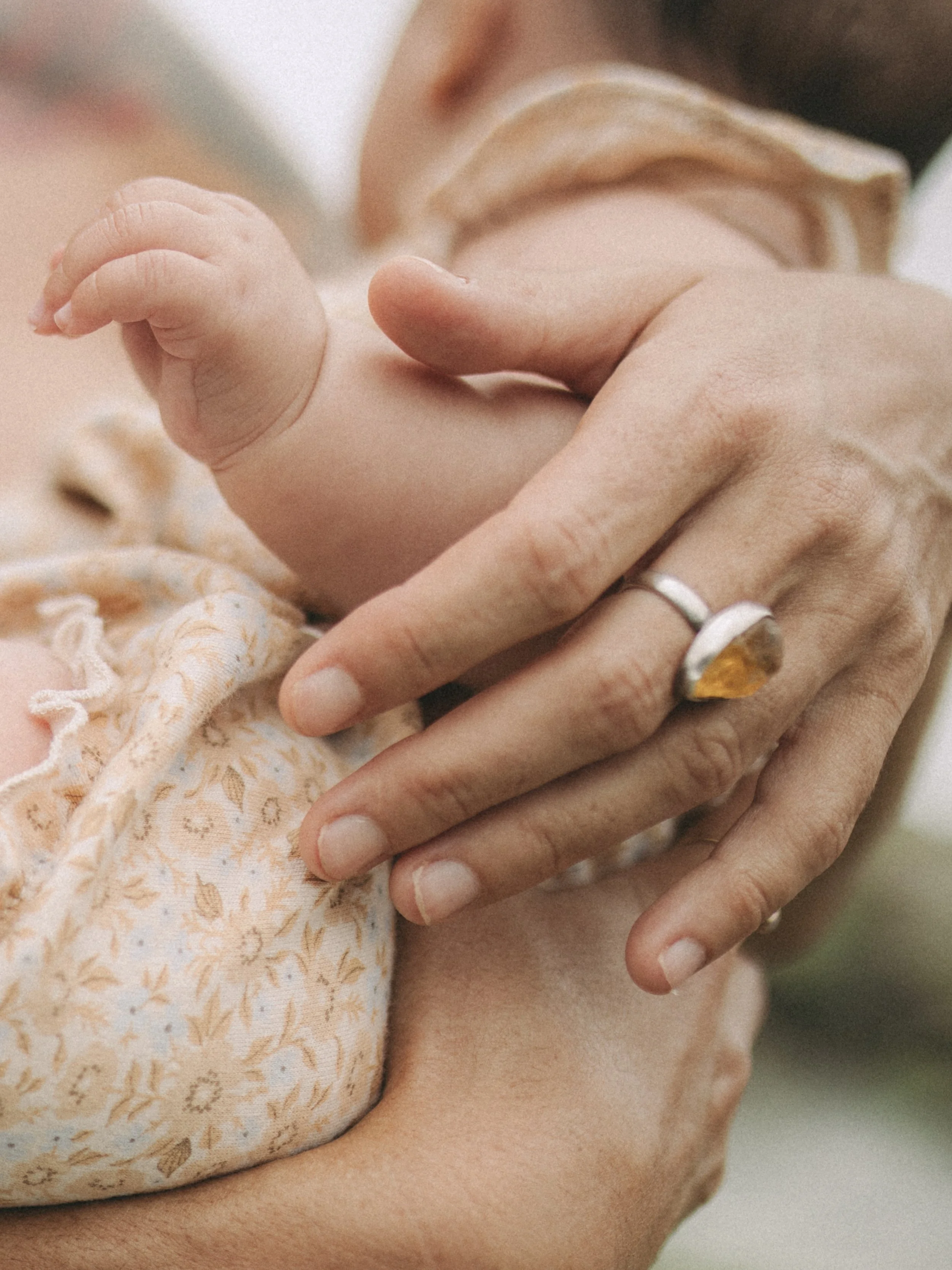 Close-up of an adult holding a baby's hand gently while caring for the baby's head and shoulder, with the baby wearing a patterned onesie.