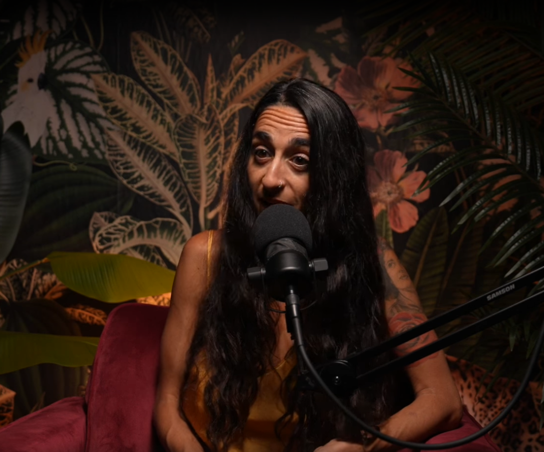 A woman with long dark hair and tattoos sitting in front of a microphone in a room with tropical plants and flower wallpaper.