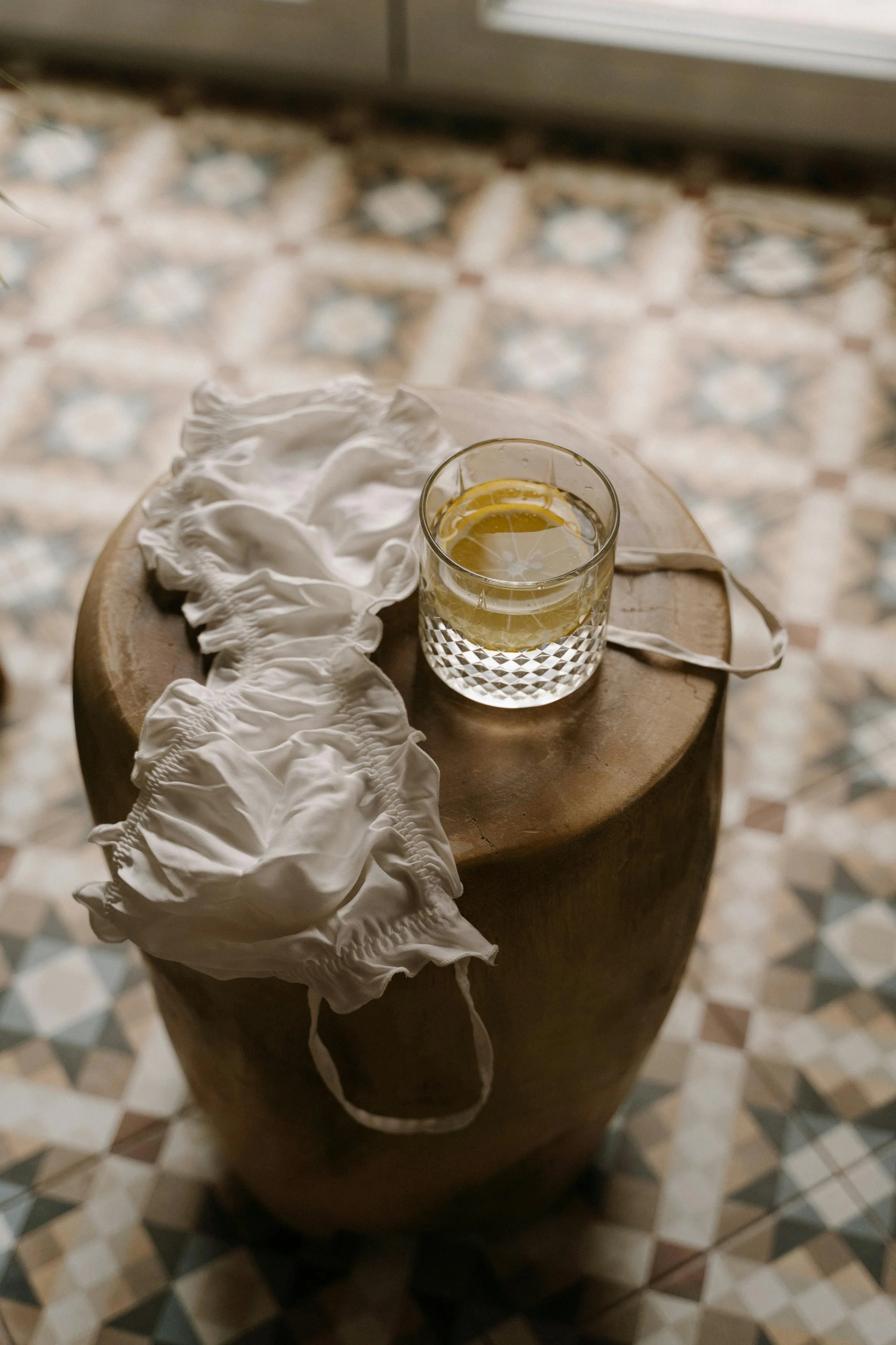 A glass of water with lemon slices on a wooden stool, next to white cloth face masks, on a patterned tile floor.