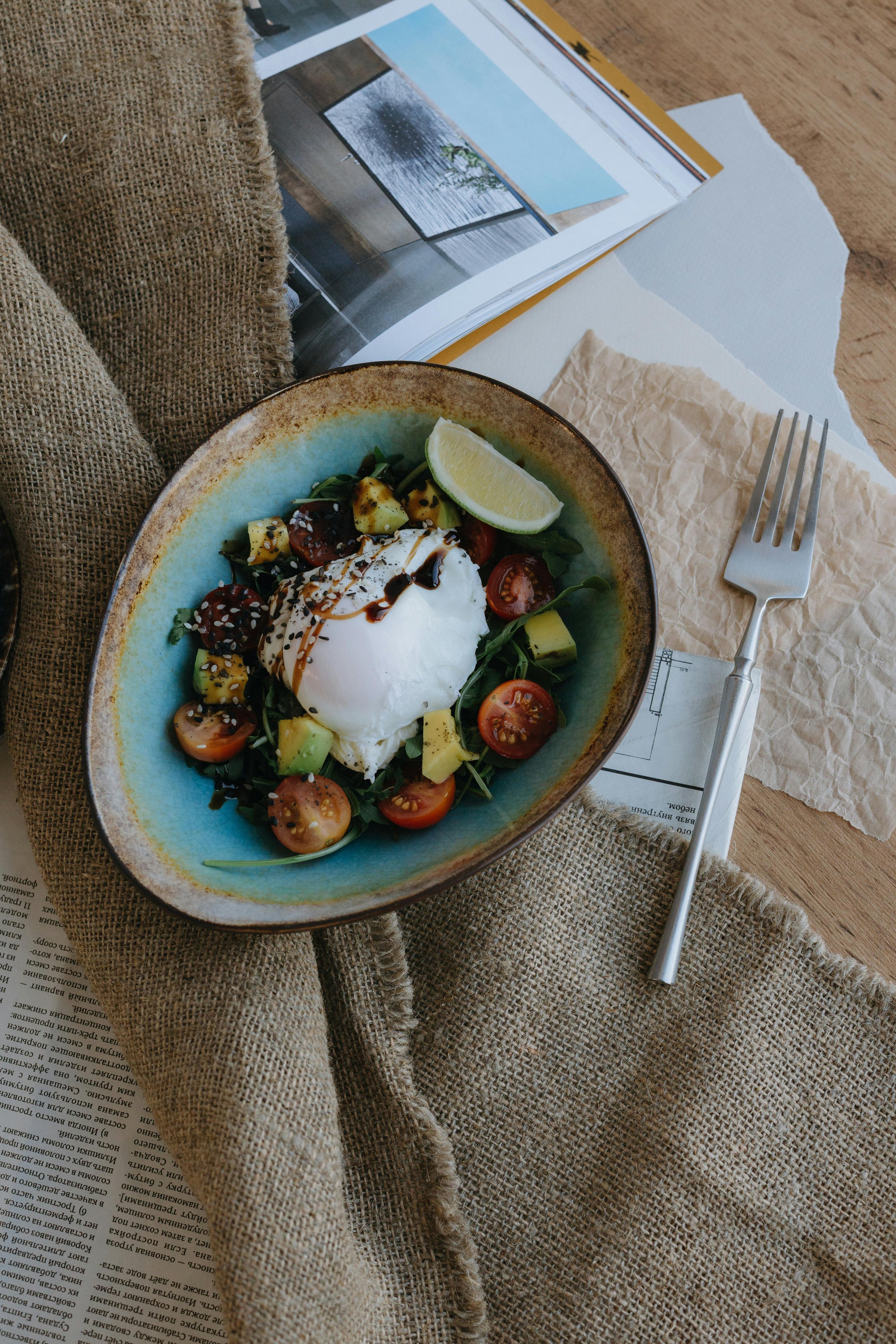 A bowl of salad with cherry tomatoes, diced avocado, and a poached egg topped with balsamic glaze, served with a wedge of lemon.