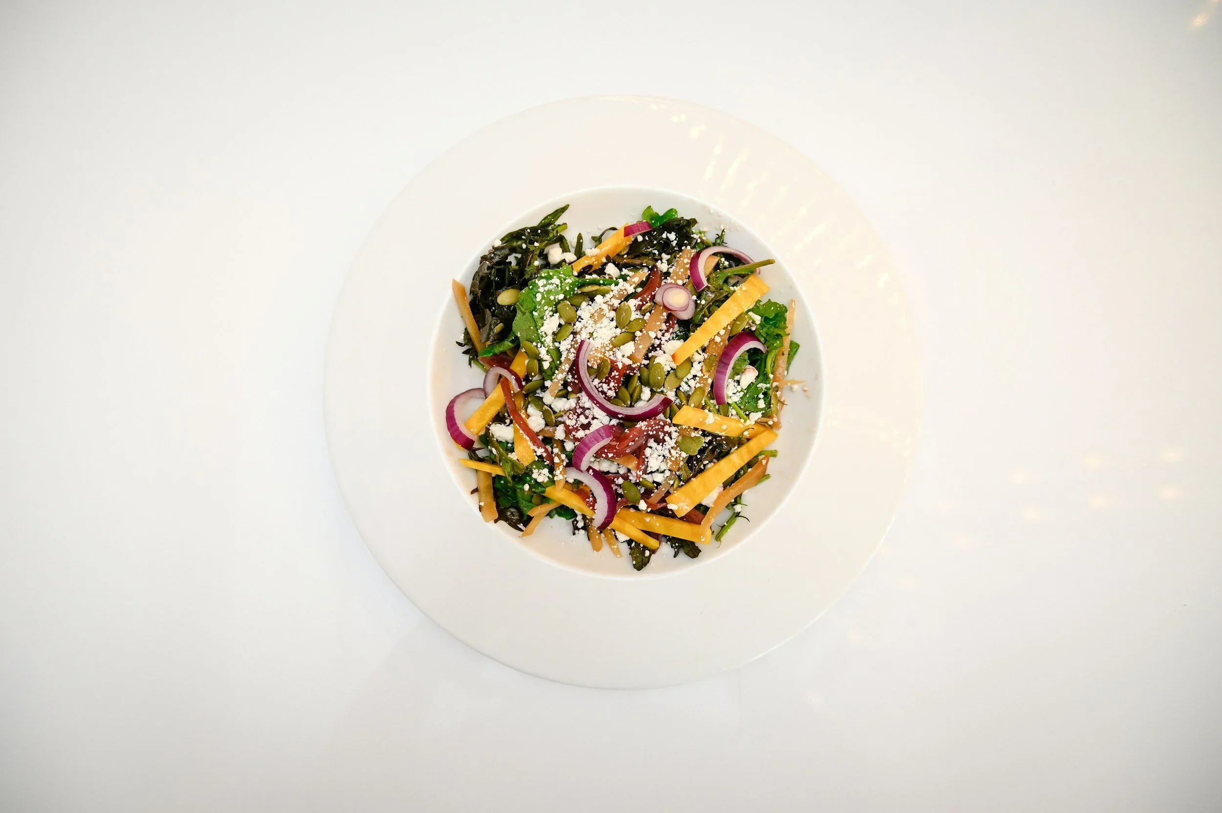 Salad with mixed greens, shredded cheese, red onions, pumpkin seeds, and shredded carrots on a white plate against a white background.