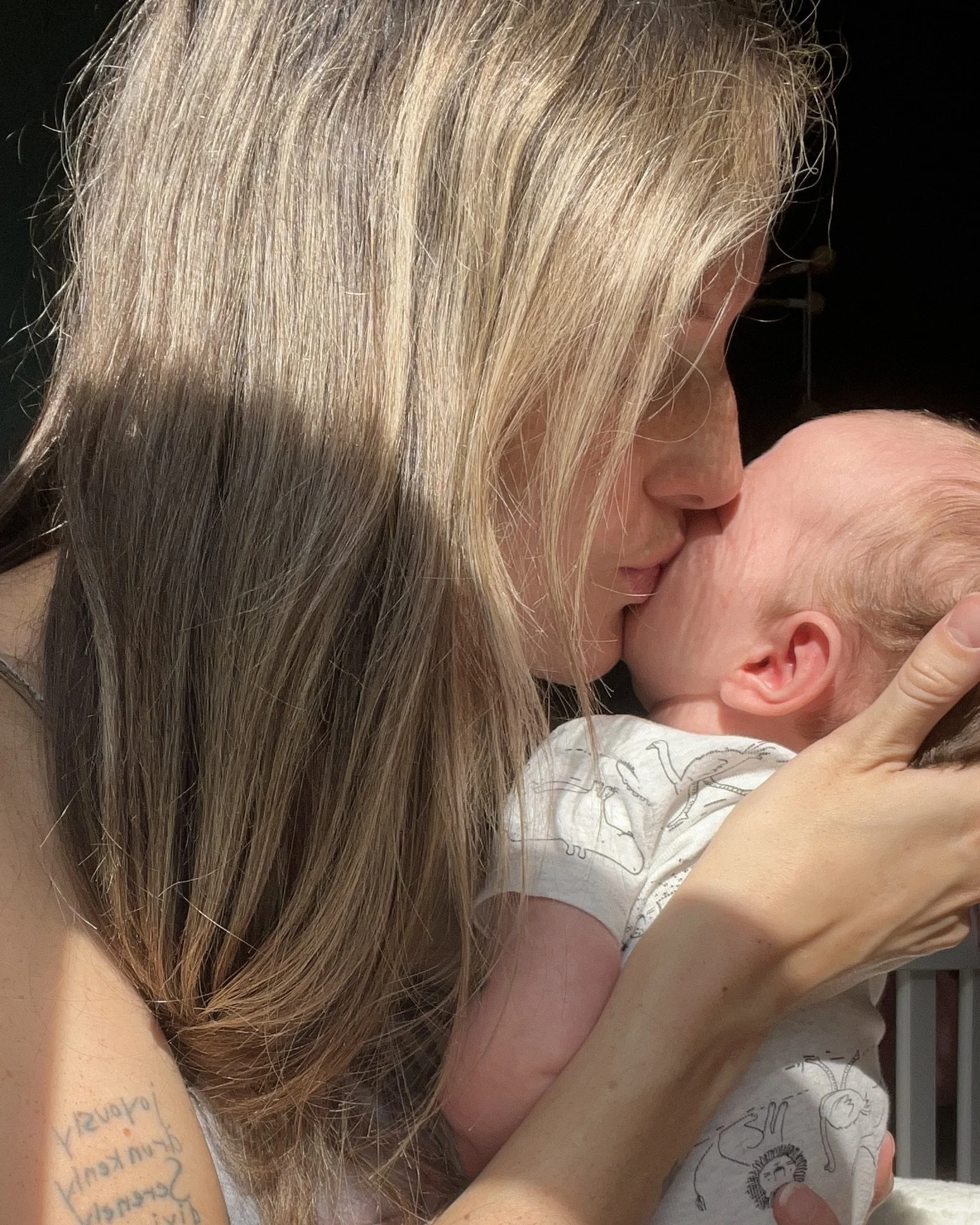 A woman with long blonde hair gently holding and kissing a newborn baby with light-colored hair.