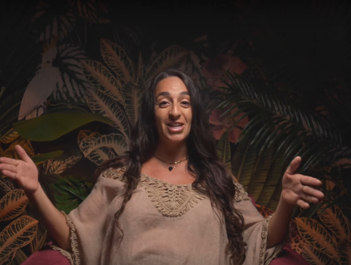 Woman with long dark hair speaking in front of tropical plant backdrop, wearing a beige blouse and a necklace.