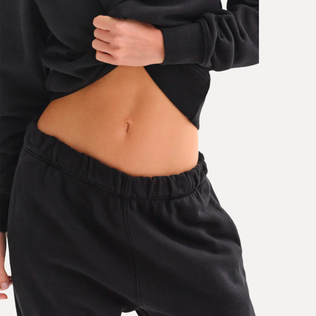 Close-up of a person wearing black sweatpants and a black hoodie, lifting the hoodie to reveal their bare midriff and navel.