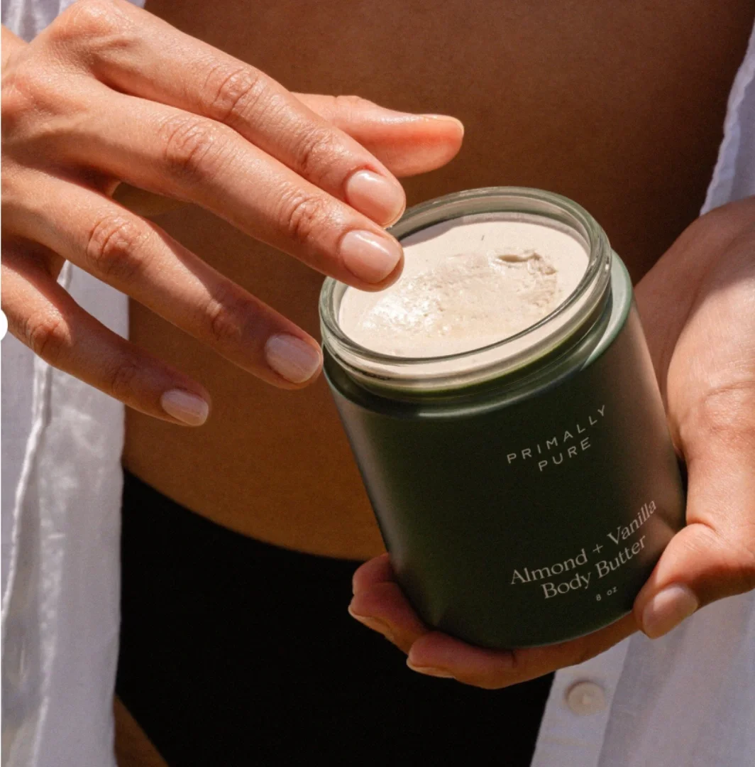 A person holding a jar of almond and vanilla body butter with the lid partially open, showing the creamy product inside.