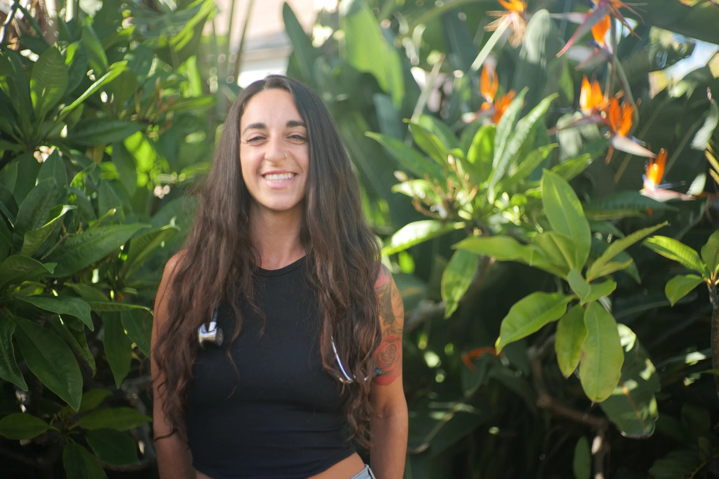 A smiling woman with long wavy hair, wearing a black sleeveless top, standing outdoors among green foliage with orange flowers.
