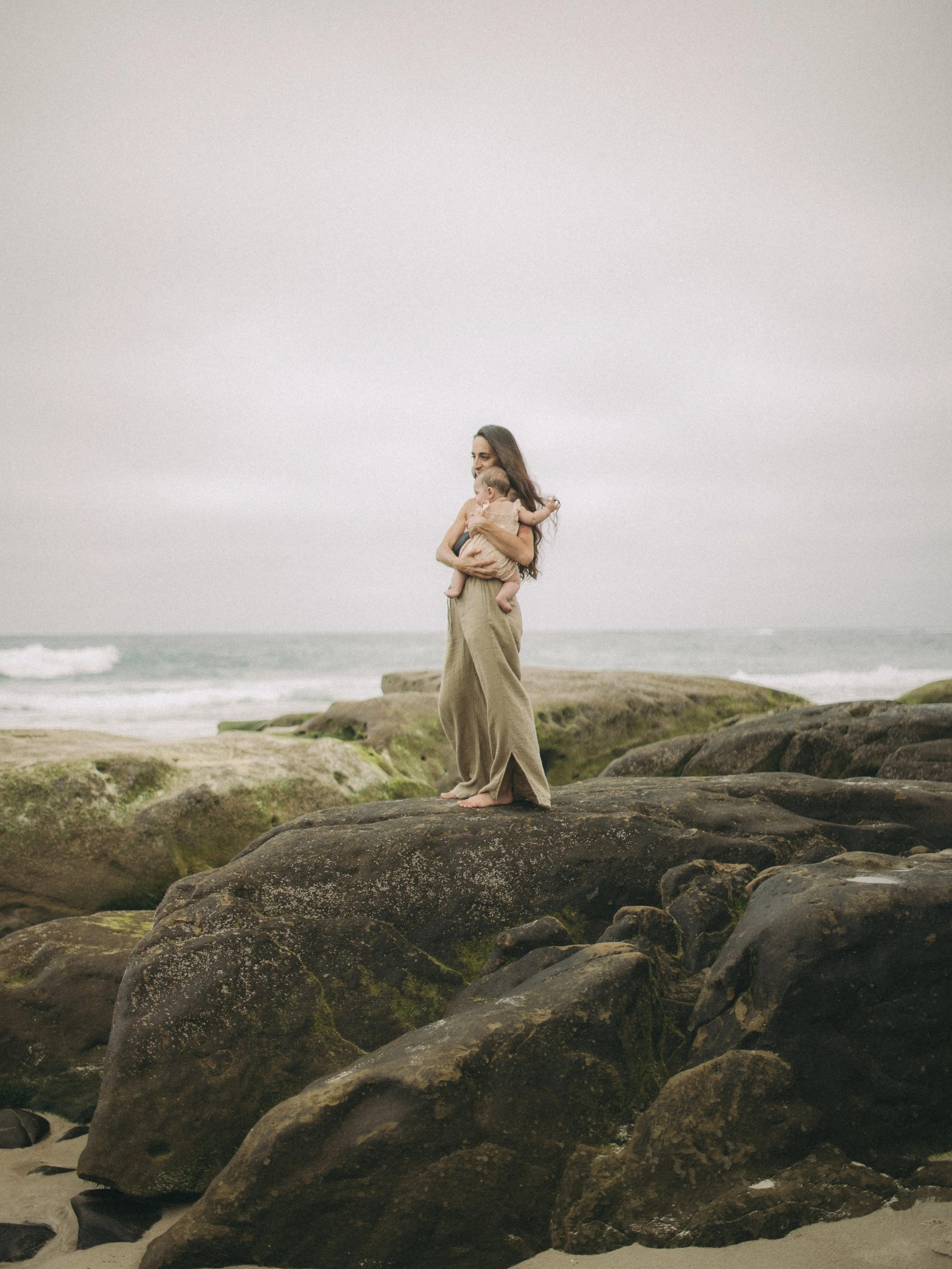 A woman holding a baby on a rocky beach with ocean waves and cloudy sky in the background.