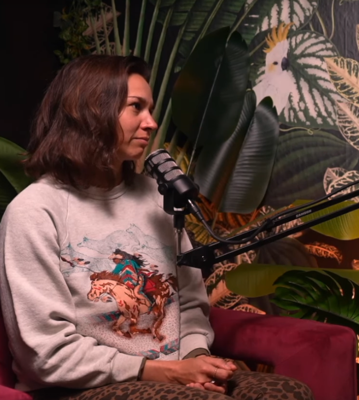 A woman with brown hair sitting in front of a microphone, against a tropical-themed background with large leaves and a bird illustration.