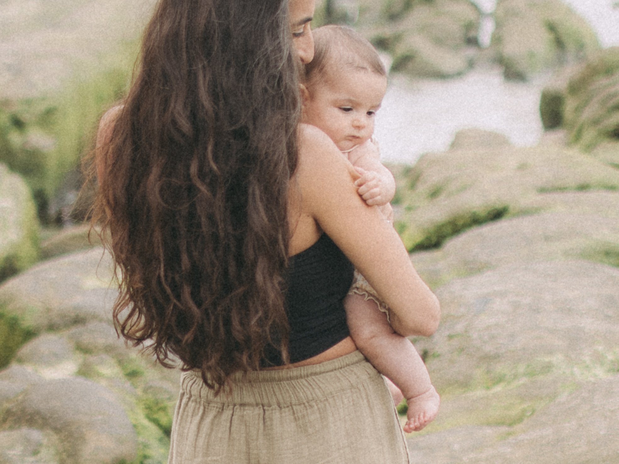 A woman with long, wavy hair holding a baby girl in an outdoor setting with rocks and greenery.
