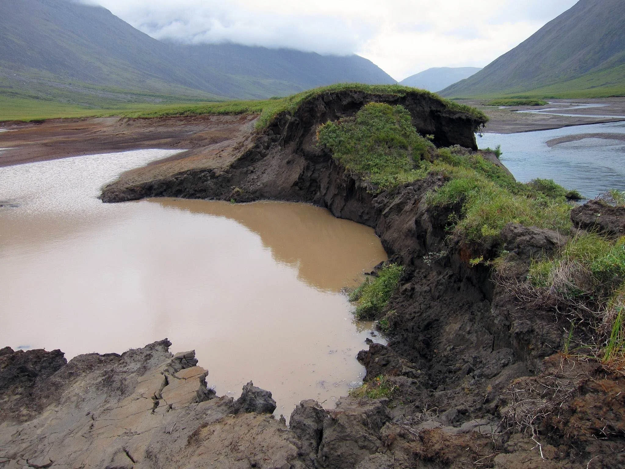 A landscape with a damaged riverbank showing exposed soil and vegetation, surrounded by mountains and cloudy sky in the background.