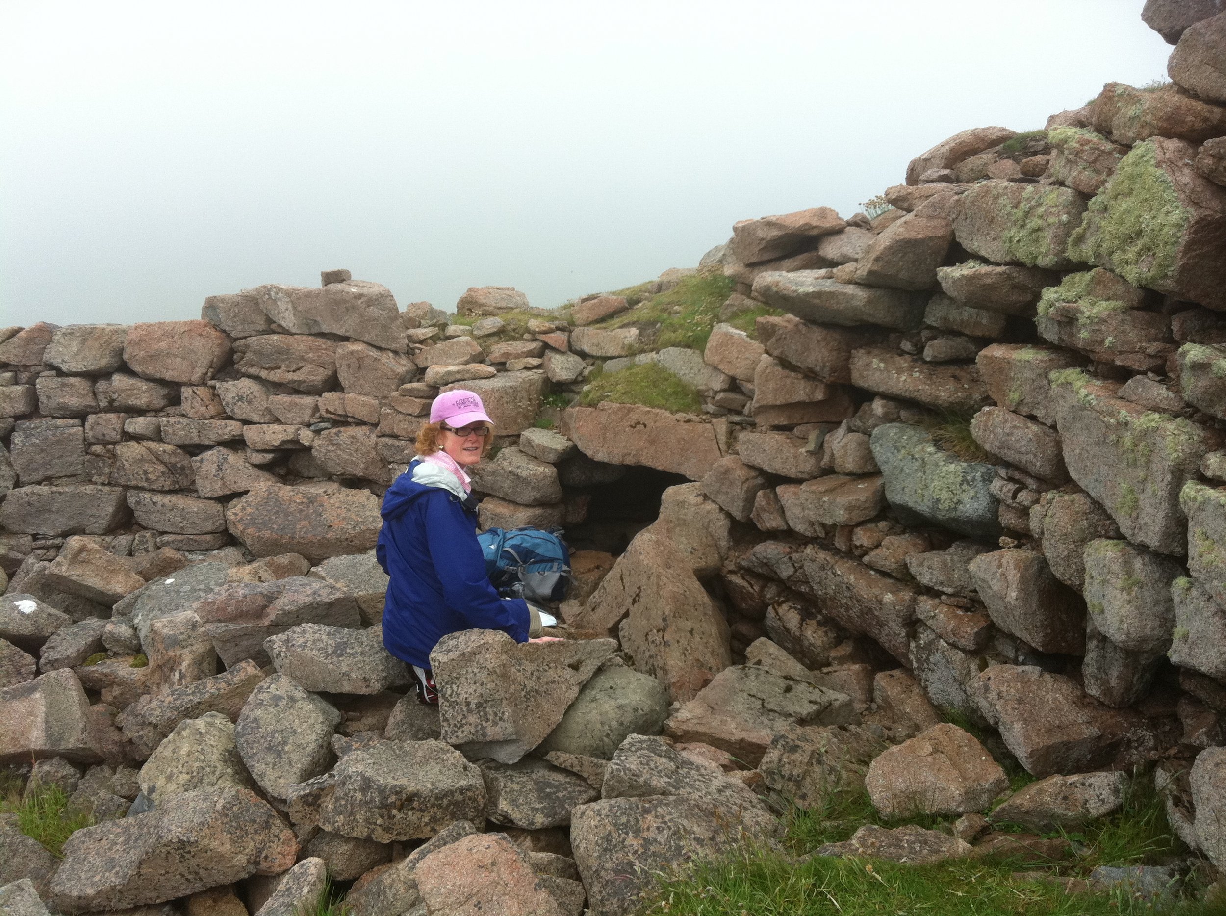 A woman in a blue jacket and pink hat smiling while sitting on rocks amid an ancient stone wall, with foggy weather and green moss on the stones.