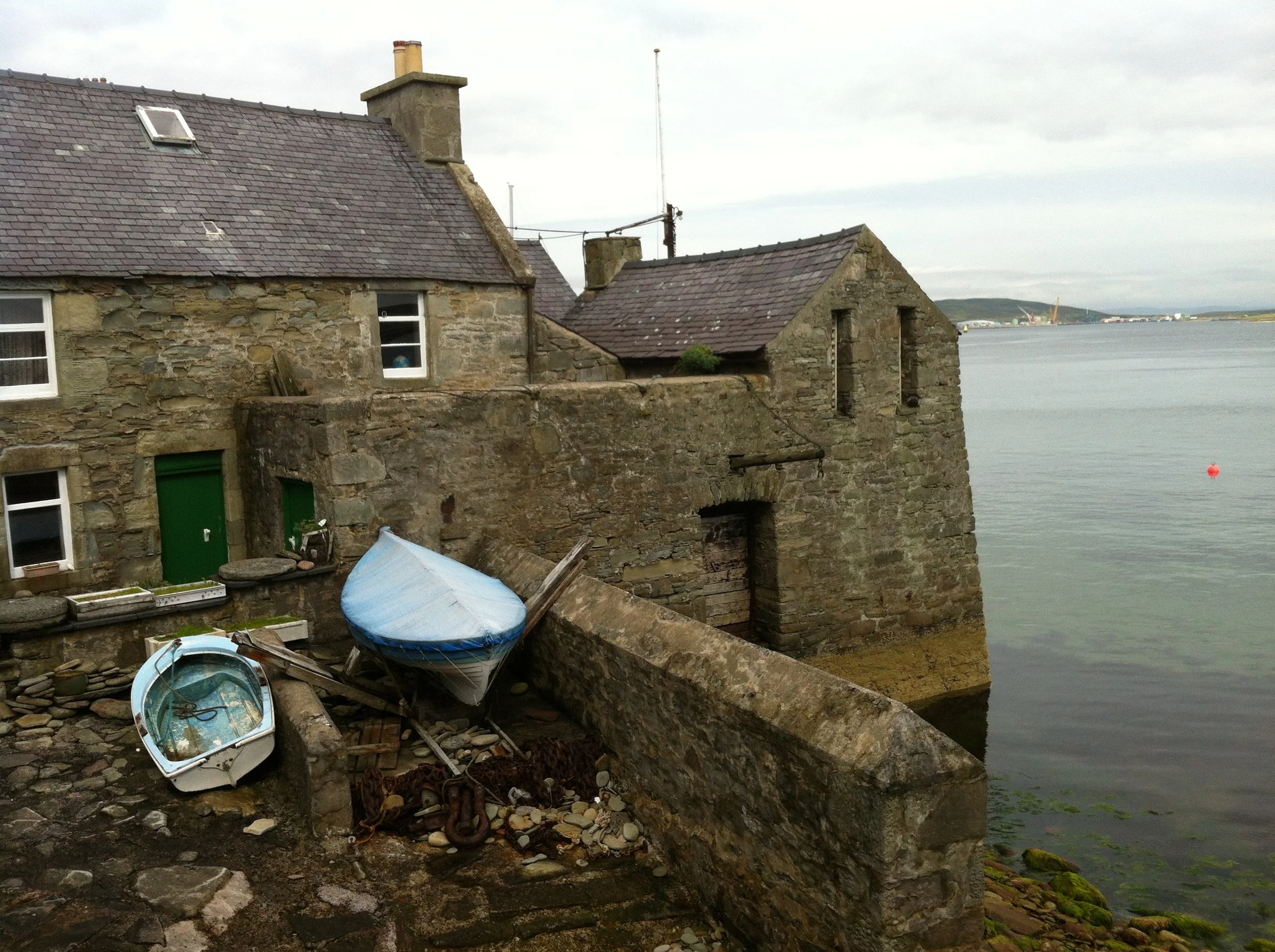 Old stone houses by the waterfront with boats overturned on the rocky shore, cloudy sky in the background.