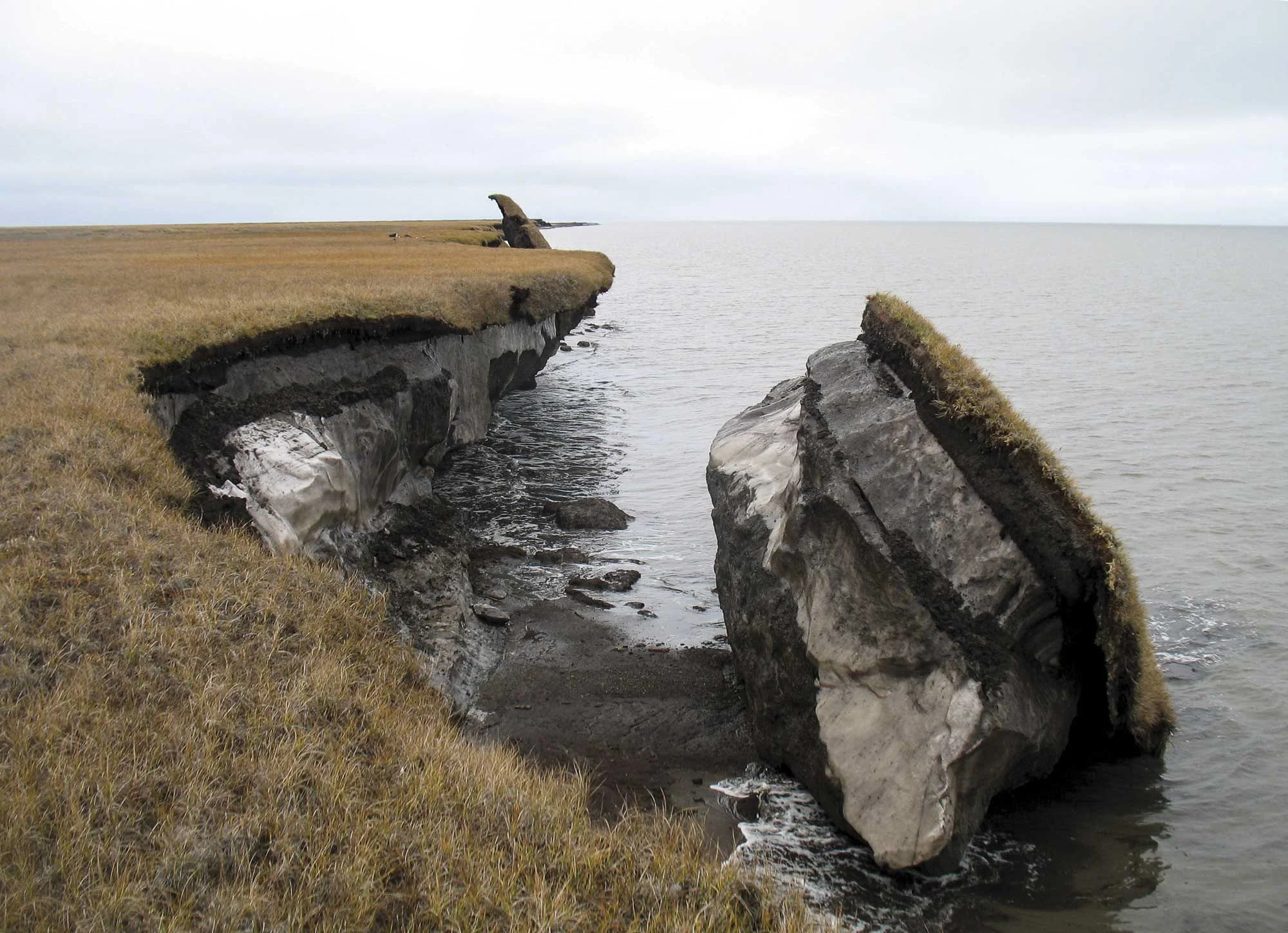 Cliffs with a large split rock formation surrounded by water and grassy terrain under an overcast sky.