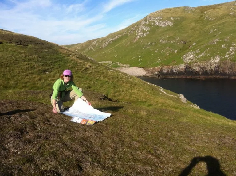 Emilie Christie Burack in a green jacket and pink hat sitting on grass with a large map in a hilly landscape near a body of water.