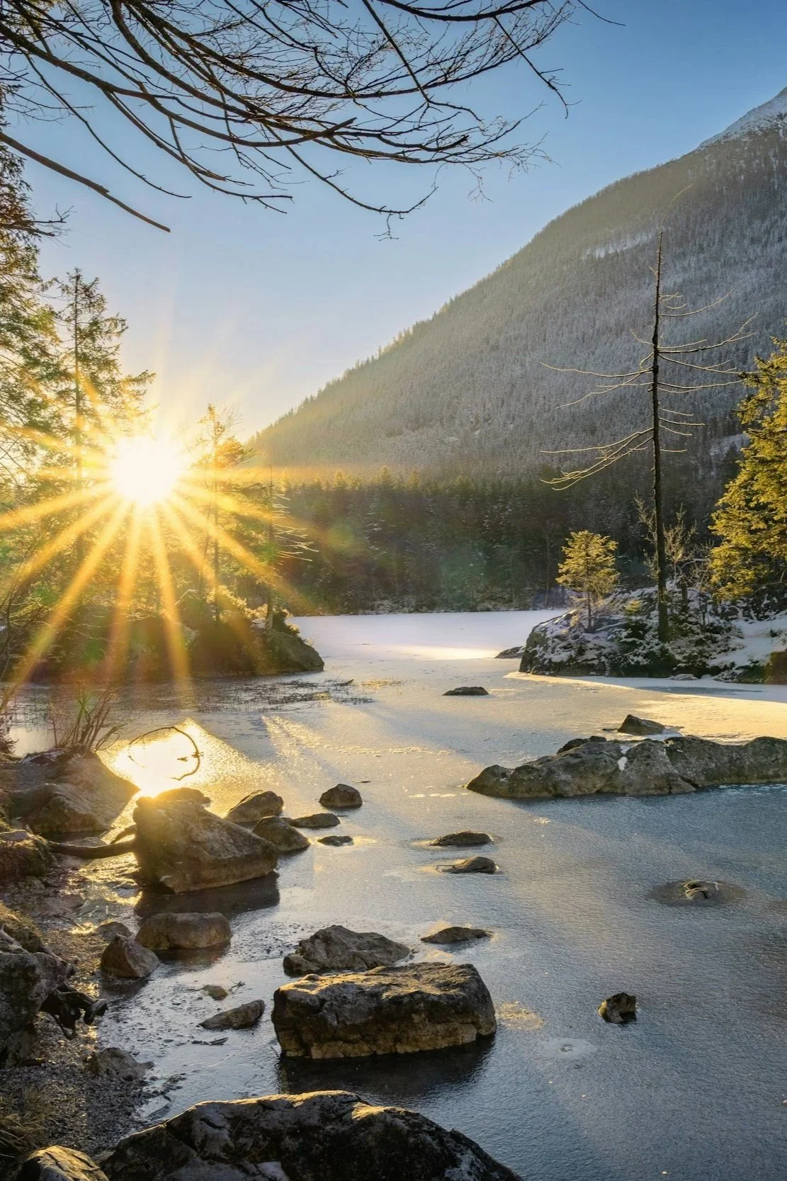 Sunrise over a forested mountain river with rocks and snow, casting a warm glow.
