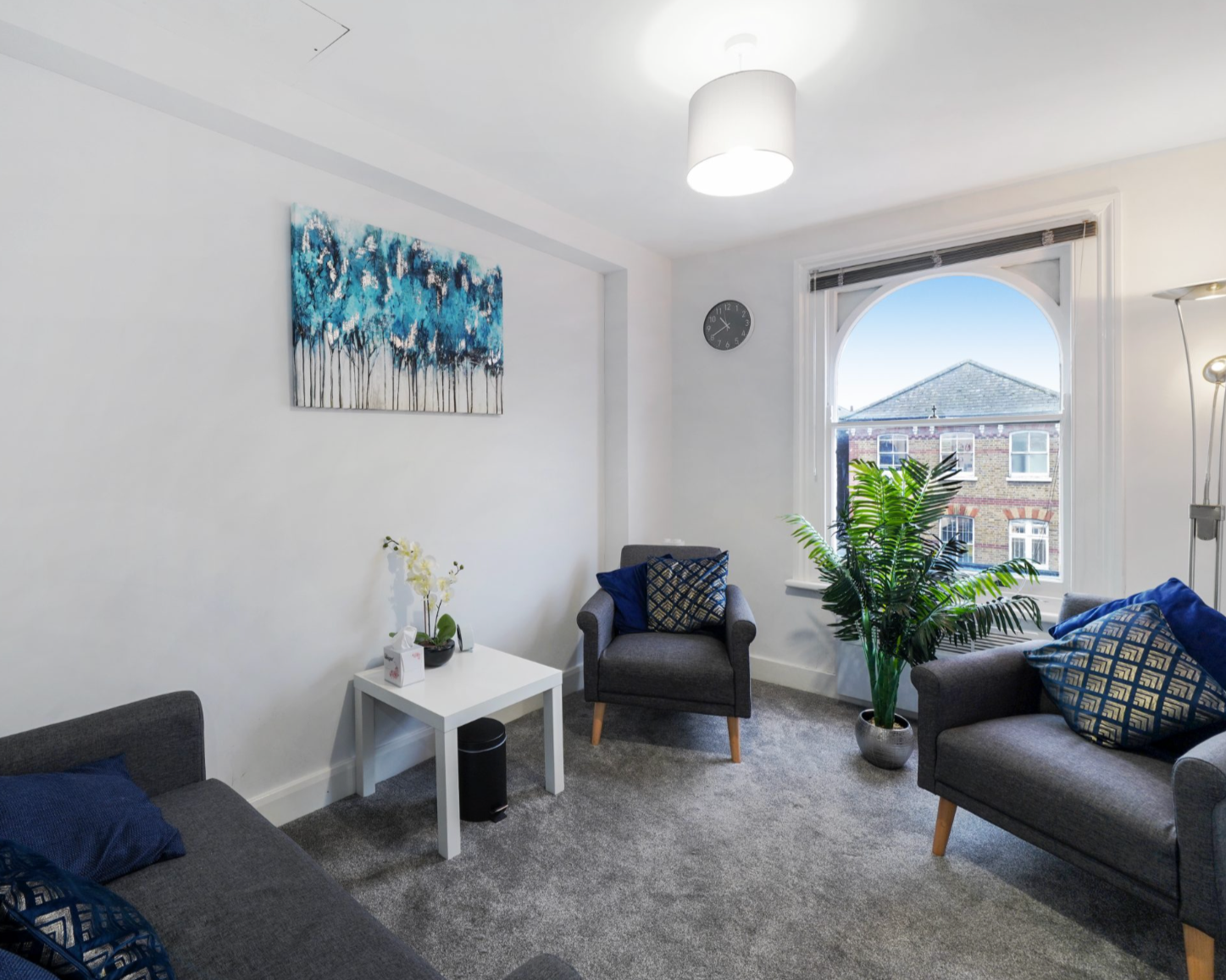 Living room with gray sofas, blue cushions, a white side table, a potted plant near a large arched window, and an abstract painting of blue trees on the wall.