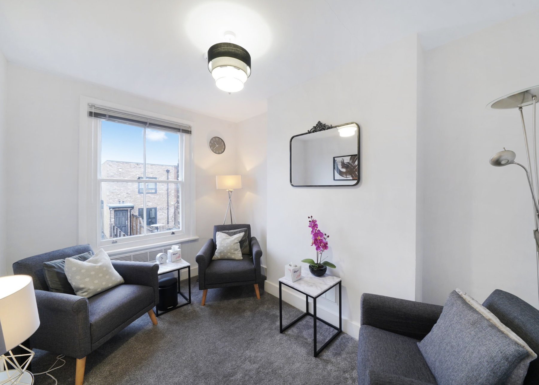Living room with three gray sofas, a window showing brick buildings outside, a small side table with a pink orchid, a round wall clock, a mirror, a standing lamp, and a ceiling light