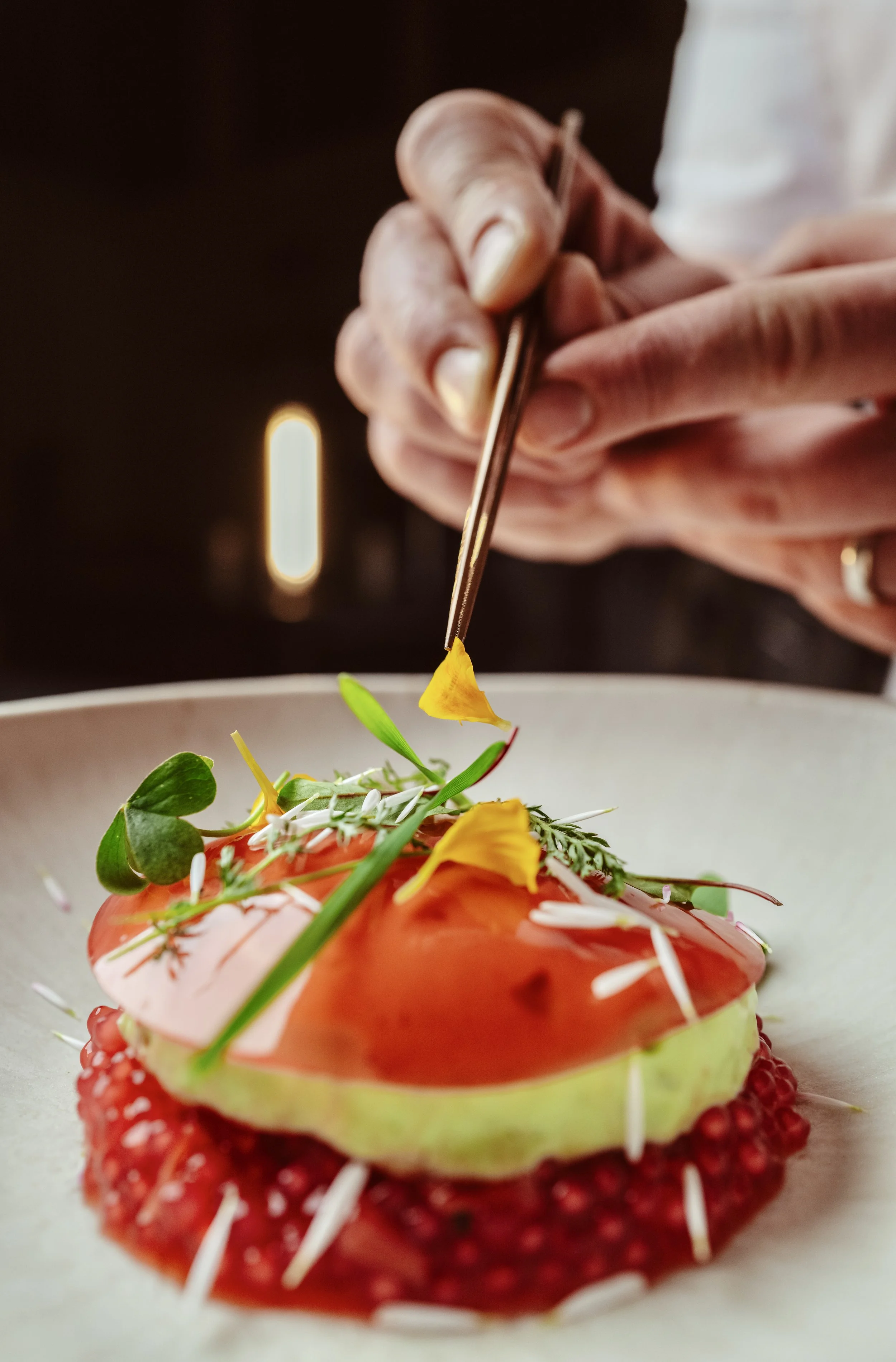 Close-up of a hand using tweezers to place edible flowers on a gourmet dish, featuring a slice of avocado, a red sauce, microgreens, and decorative garnishes on a white plate.